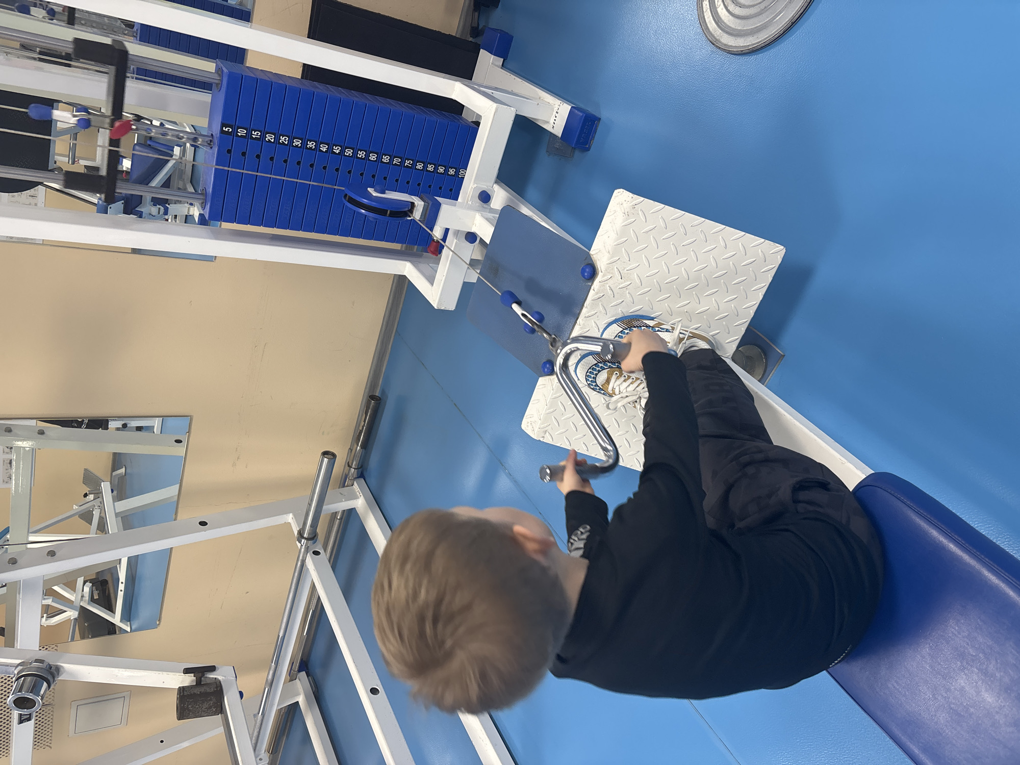 A young boy is sitting on a leg press machine in a gym. The gym has blue flooring and white exercise equipment. The machine has blue weight plates stacked on the side. The boy is wearing black pants and a black long-sleeved shirt, with white sneakers. He is grasping the handles of the leg press. The location is Chaikovsky, Russia, based on context.