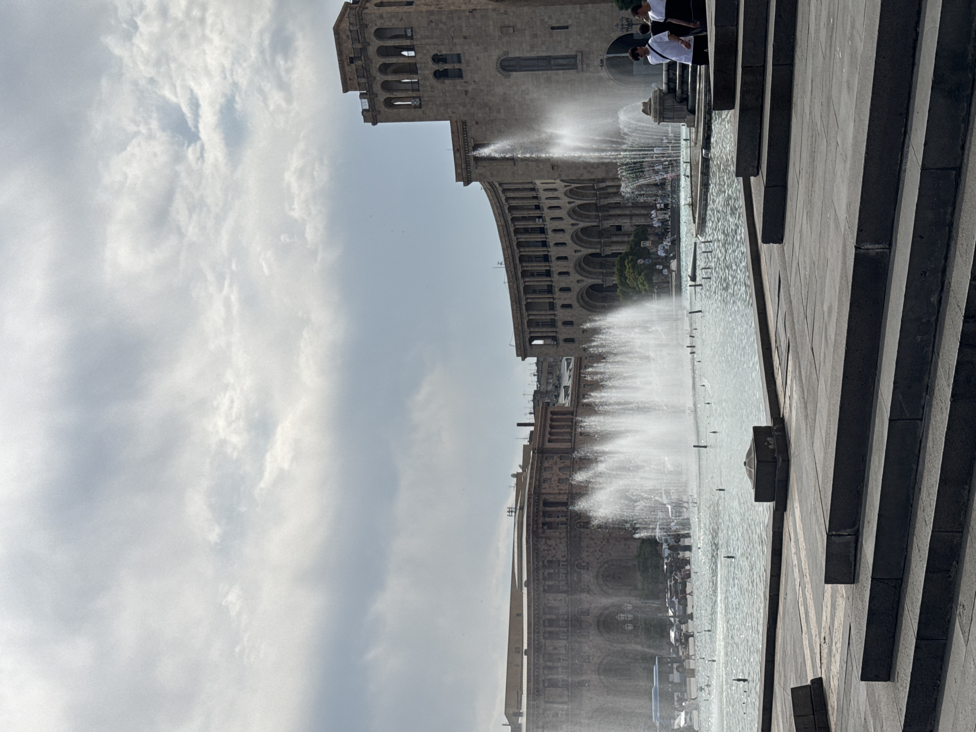 The image shows a public square in Yerevan, Armenia, featuring a large fountain in the foreground and several large stone buildings in the background. The fountain is active, with water shooting up into the air. Several people are visible, some standing near the fountain and others walking in the background. The architecture of the buildings suggests they are of historical or cultural significance. The sky is overcast, with clouds present. The overall scene depicts a lively urban environment with a prominent water feature.