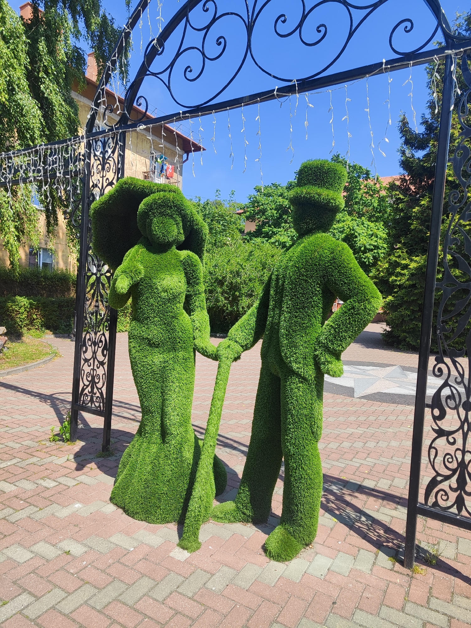 Two topiary figures, a woman in a long dress and a man in a hat and suit, stand on a brick-paved walkway in front of a black ornate archway decorated with string lights. The figures are made of green artificial grass. The woman's left hand rests on her hip, and she holds a long green pole with her right hand. The man's left hand is on his hip, and his right hand also holds the green pole. Behind the figures, there are trees and a building with a red-tiled roof. The scene is illuminated by bright sunlight, casting shadows on the ground. The location is the Kurortny Prospekt in Pionersky, Russia.