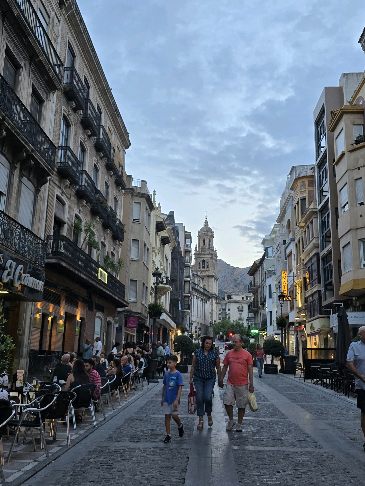 The image depicts a street scene in Jaén, Spain. On the left side of the street, there is a building with balconies and what appears to be a restaurant with outdoor seating. The restaurant has tables and chairs set up along the sidewalk, and several people are seated at the tables. On the right side of the street, there are more buildings with shops and businesses. A church tower is visible in the background. Several people are walking on the street, including a family holding hands. The sky is cloudy.