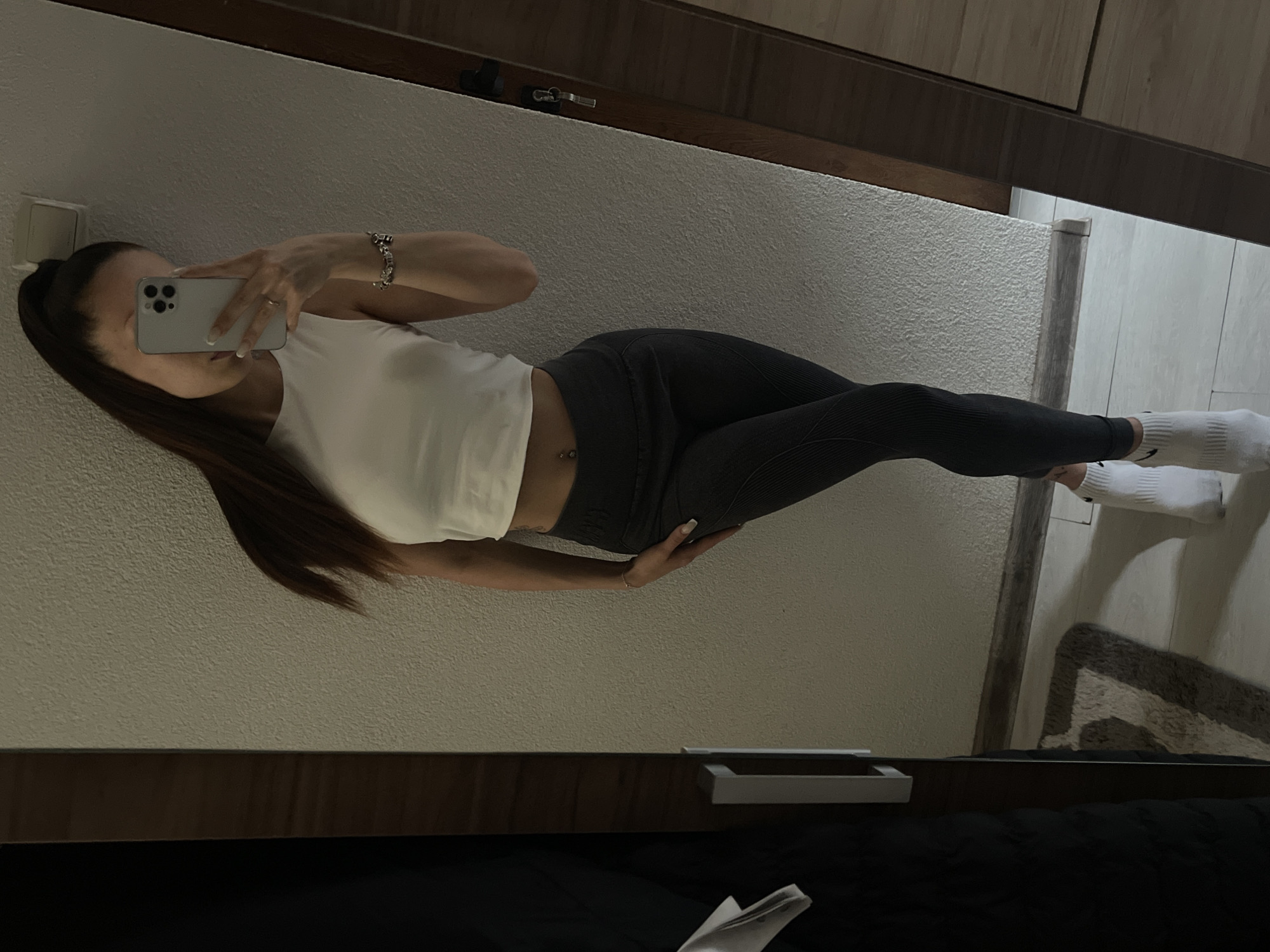 A young woman is taking a mirror selfie in what appears to be a bedroom or dressing area. She is standing and leaning against a textured wall. She is wearing a white crop top, dark gray ribbed leggings, and white socks. Her long, dark hair is pulled back into a high ponytail. She is holding a smartphone in her right hand, using it to capture the reflection. A bracelet adorns her left wrist, and a belly button piercing is visible. To the left of the frame, a portion of a dark closet or wardrobe is visible, with a white garment hanging on the inside. The floor is tiled, and a rug with a geometric pattern is partially visible in the lower left corner. The right side of the image shows a mirror and a doorframe. The lighting is dim, creating shadows. There is no indication of the location being in a city.