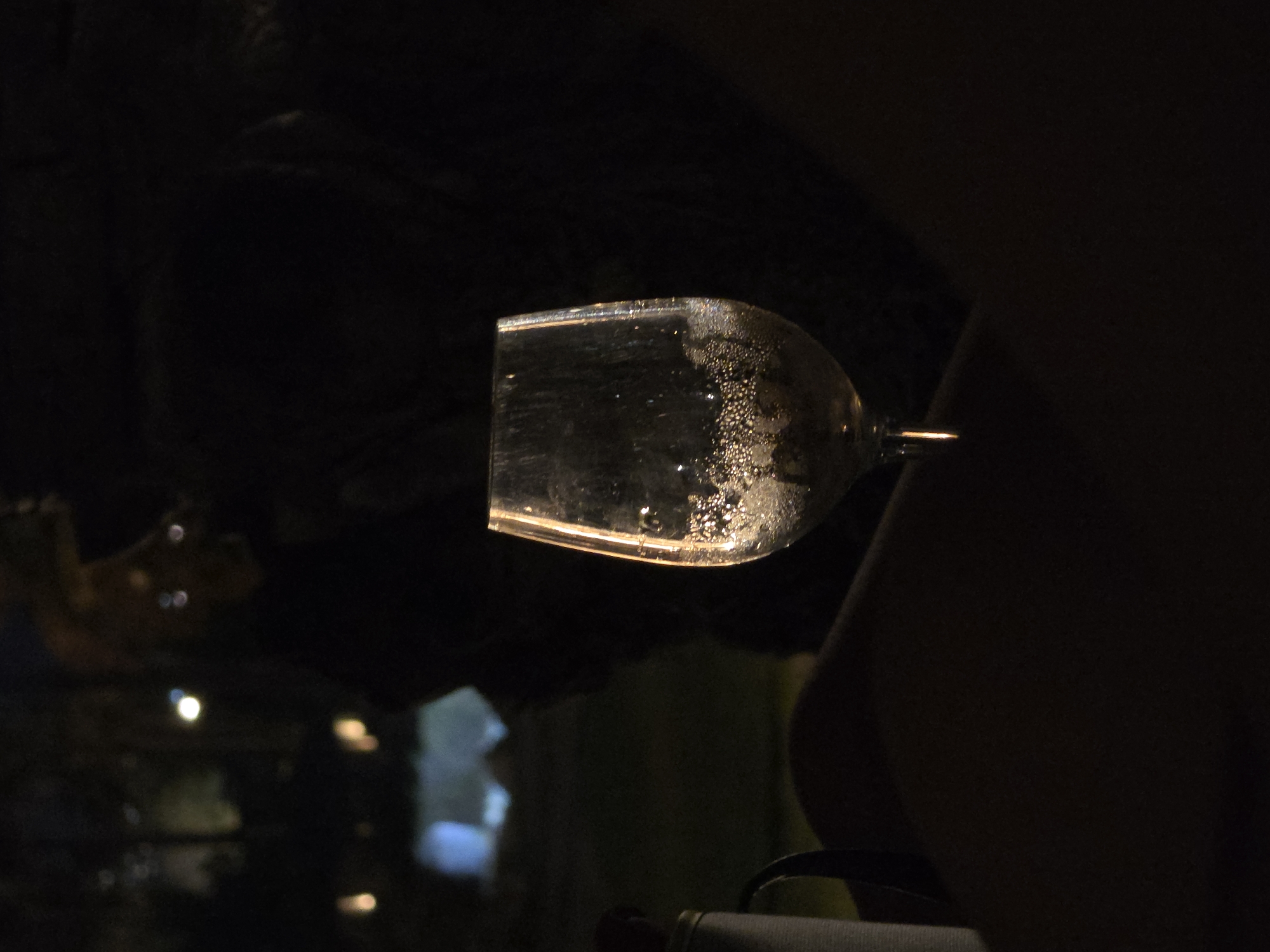 A wine glass filled with a clear liquid, possibly wine or water, sits on a dark surface, possibly a lap or table, in the foreground of the image. Bubbles are visible within the liquid, clinging to the sides of the glass. The glass is illuminated from behind, creating a bright rim and highlighting the condensation on its exterior. To the left of the glass, a cylindrical object with a handle, possibly a thermos or bottle, is partially visible. The background is dark and indistinct, with faint lights suggesting an outdoor setting at night, possibly in the Hlorakas Community of Cyprus. There are no discernible people in the image.