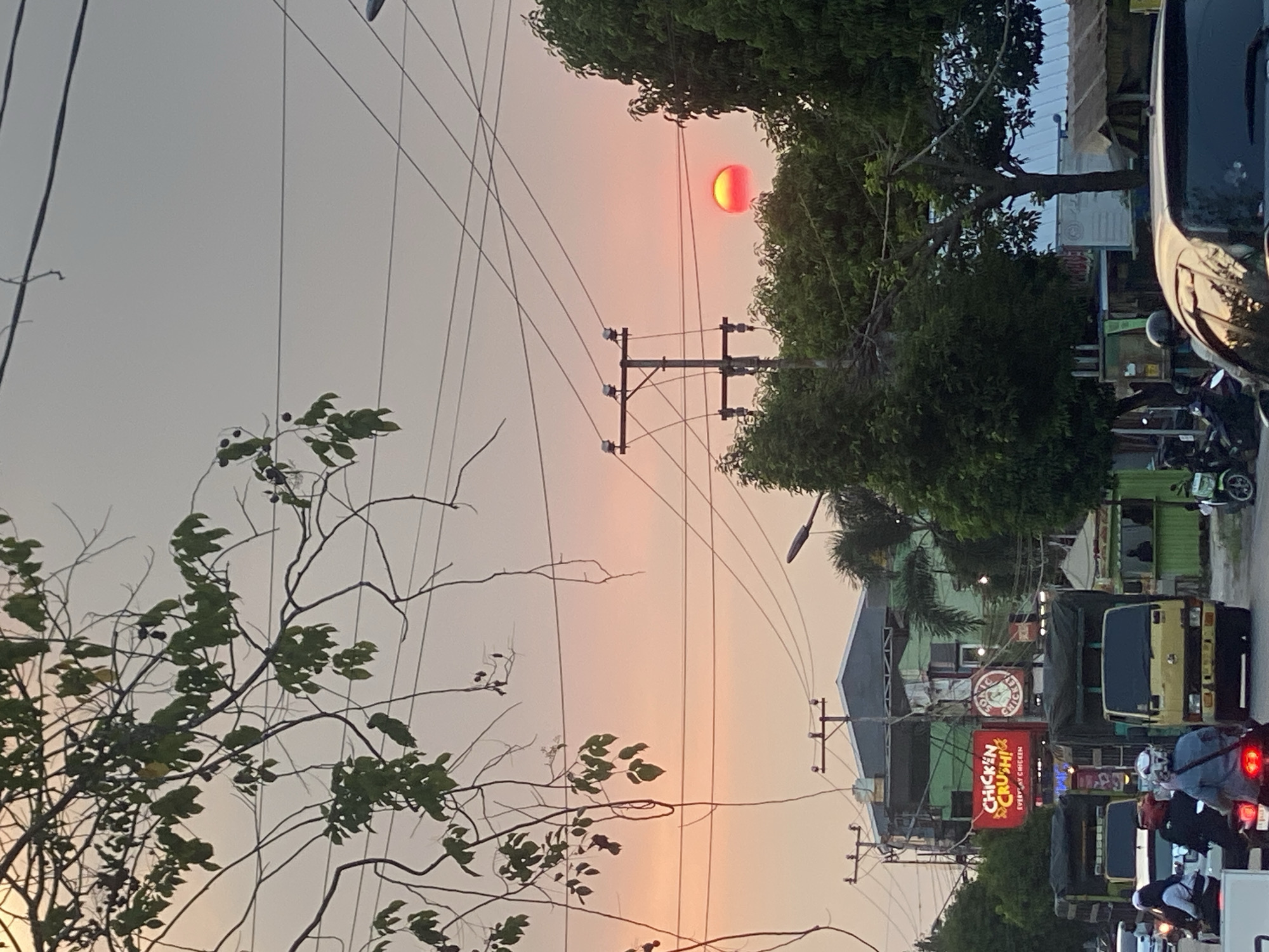 The image captures a street scene in Banjarbaru, Indonesia, during sunset. The sky is a gradient of soft orange and pink hues. In the middle ground, a vibrant red sun is visible, partially obscured by trees and utility poles. The street is busy with traffic, including several trucks, motorcycles, and cars. People on motorcycles are a prominent feature of the foreground. Buildings line the street, with some displaying illuminated signs, including one for "CHICKEN CRUSH!". Overhead, a network of power lines crisscrosses the sky, adding a sense of urban infrastructure to the natural beauty of the sunset. Lush green trees are interspersed throughout the scene, creating a contrast with the man-made elements. The overall atmosphere is one of a bustling city transitioning into evening.