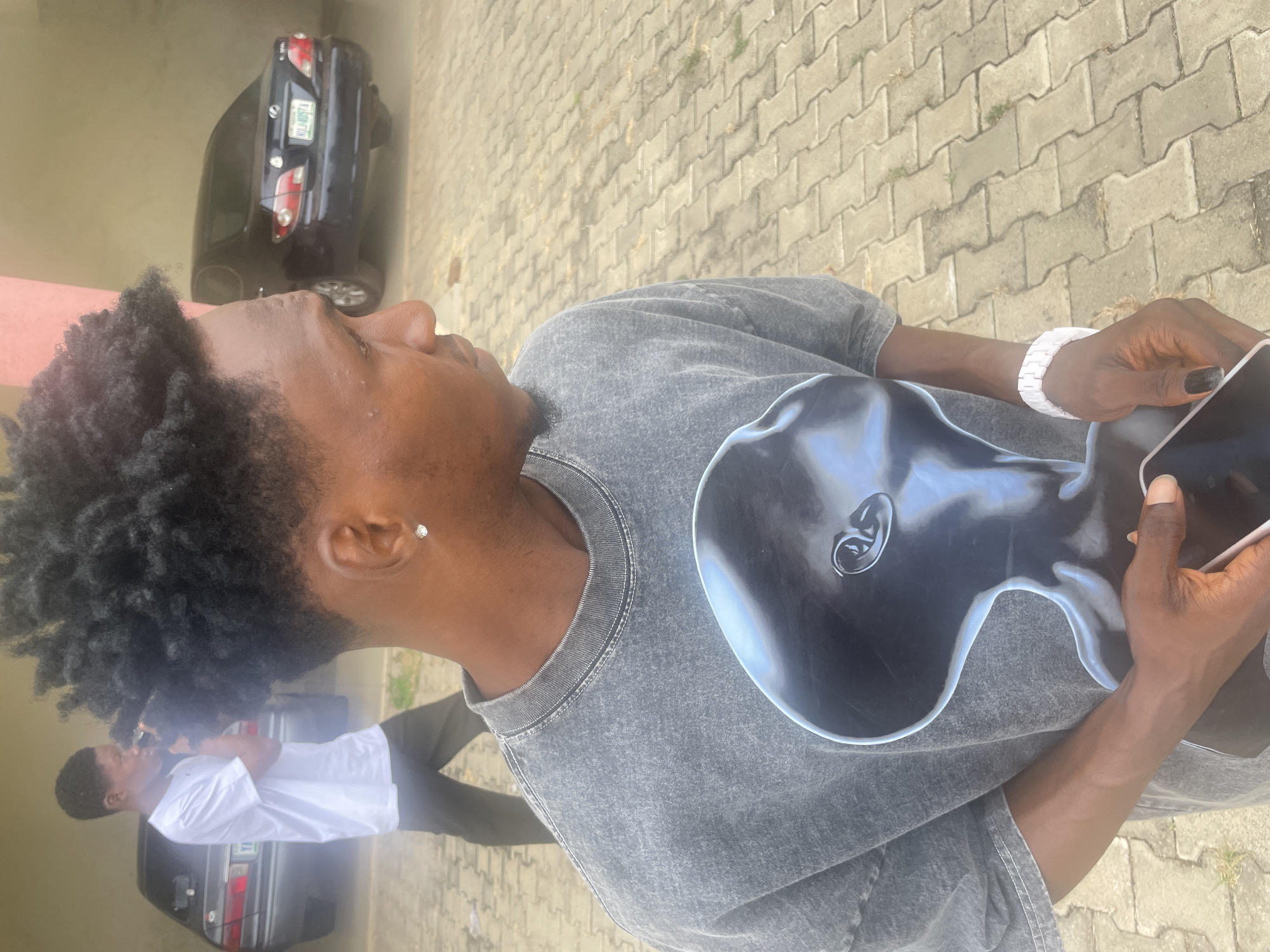 A person is holding a cell phone and standing on a gray-paved surface, possibly a sidewalk or driveway. The person wears a gray t-shirt, a white bracelet, and a small stud earring. The background features a black car, a building with a pink wall, and another person wearing a white shirt and dark pants standing beside a light-colored car. The location is likely somewhere in Lokoja, Nigeria.