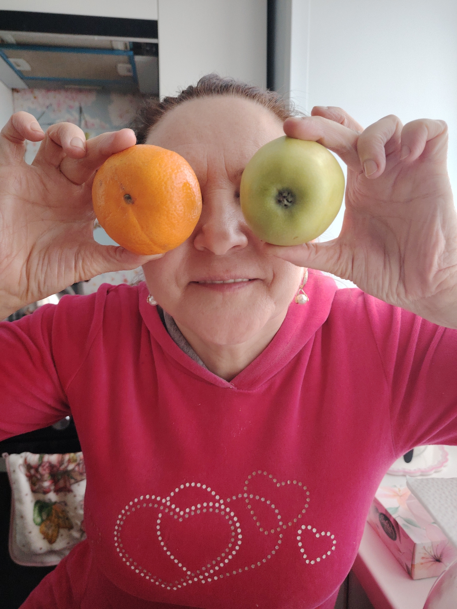 A woman is holding an orange in her left hand and a green apple in her right, positioning them in front of her eyes. She is wearing a red hooded sweatshirt with heart designs on the front. The background includes a kitchen setting with white cabinets. Location cannot be determined from the image.
