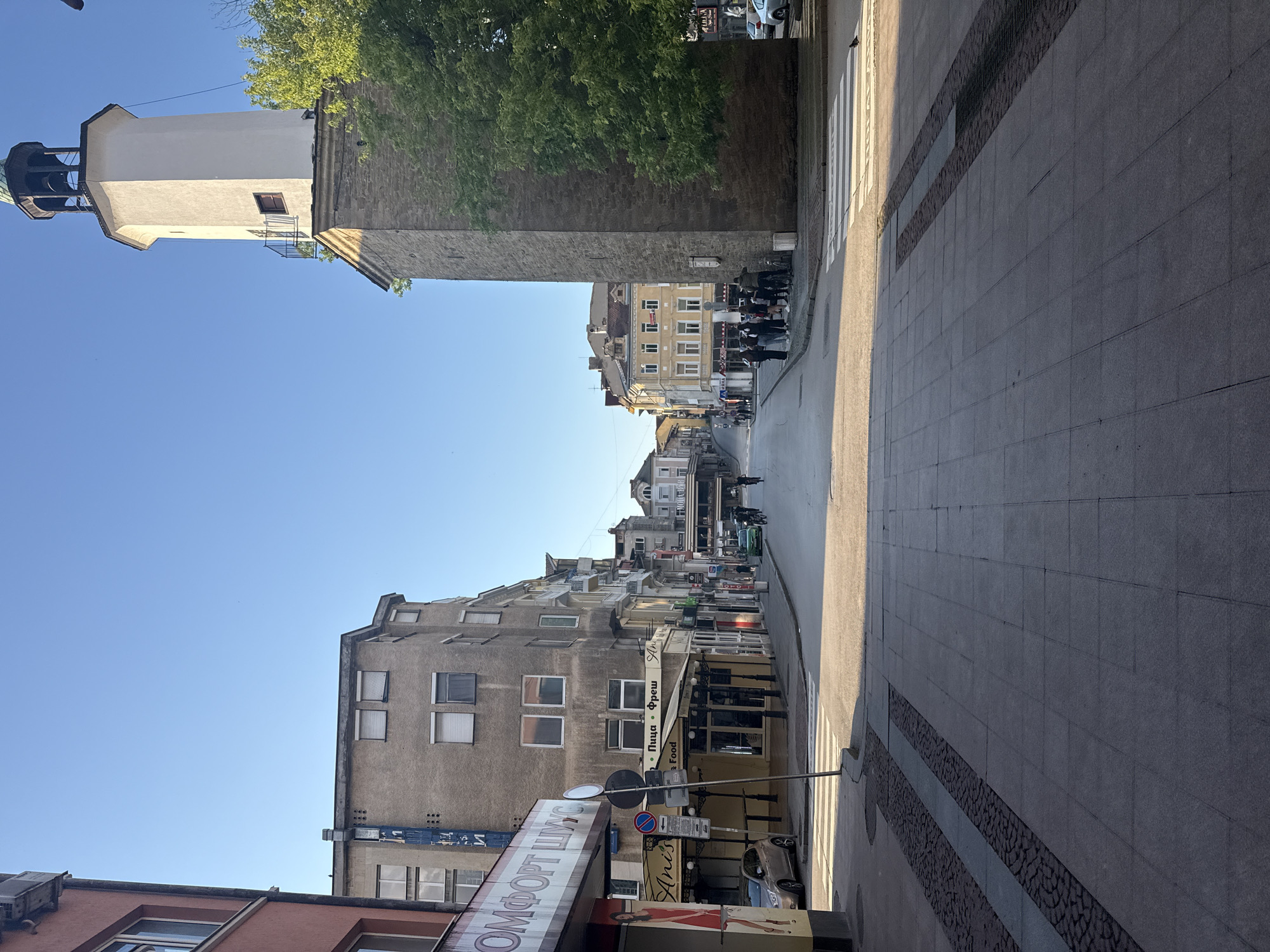 The image shows a street scene in Gabrovo, Bulgaria. A tall, square tower, possibly a clock tower or bell tower, stands to the left side of the frame, partially obscured by a tree. The street stretches into the distance, lined with buildings of varying heights and architectural styles. Some buildings have visible storefronts with signs in Cyrillic script. A zebra crossing marks a pedestrian area on the right side of the image. The sky is clear and blue. People are visible as blurry shapes in the distance walking along the street. Cars parked on the side of the street are partially visible. The ground is paved with grey stone slabs.