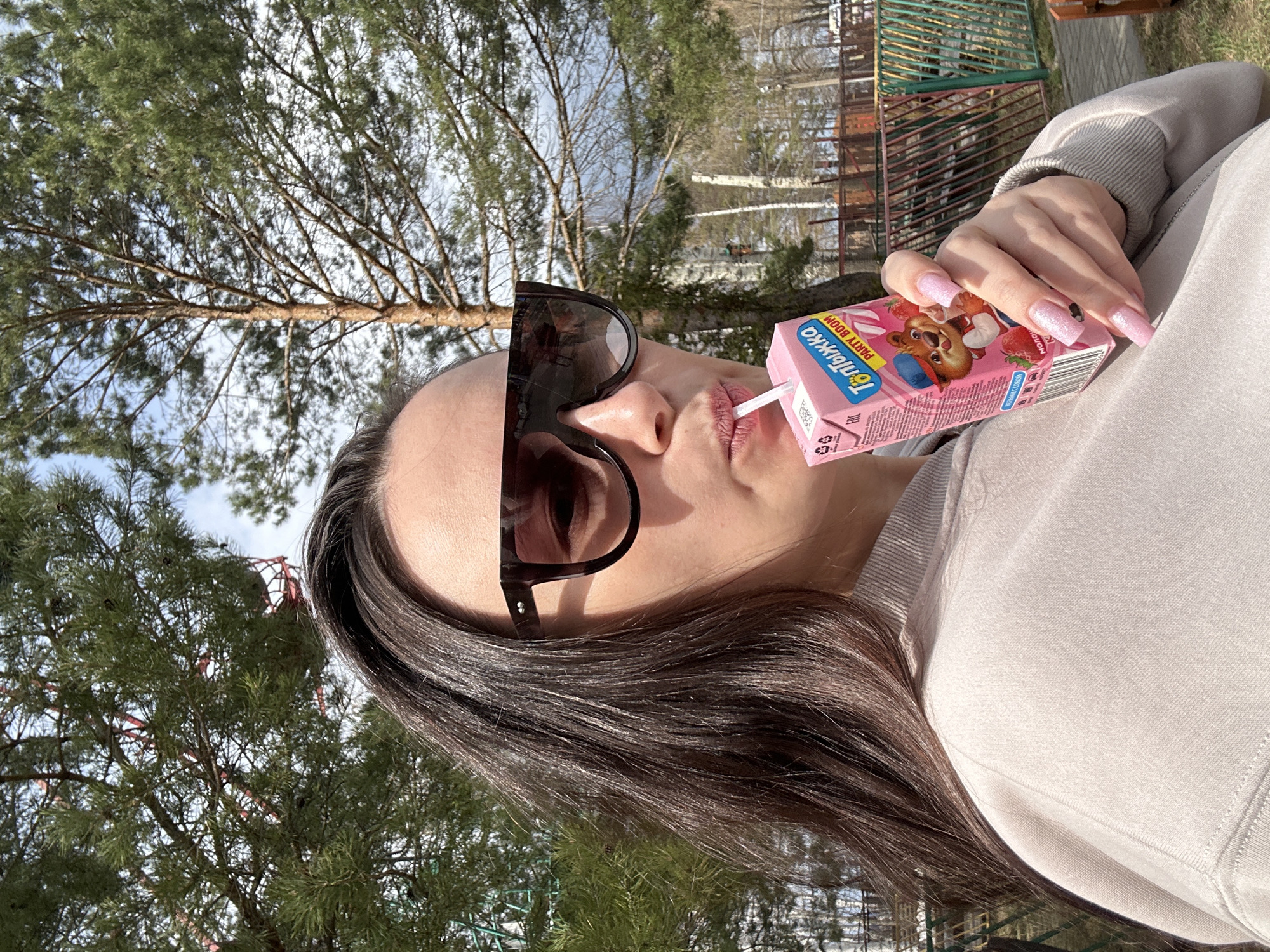 A woman wearing sunglasses is drinking a pink juice box with a straw. She is outside with trees in the background. The location is Chaikovsky, Russia.