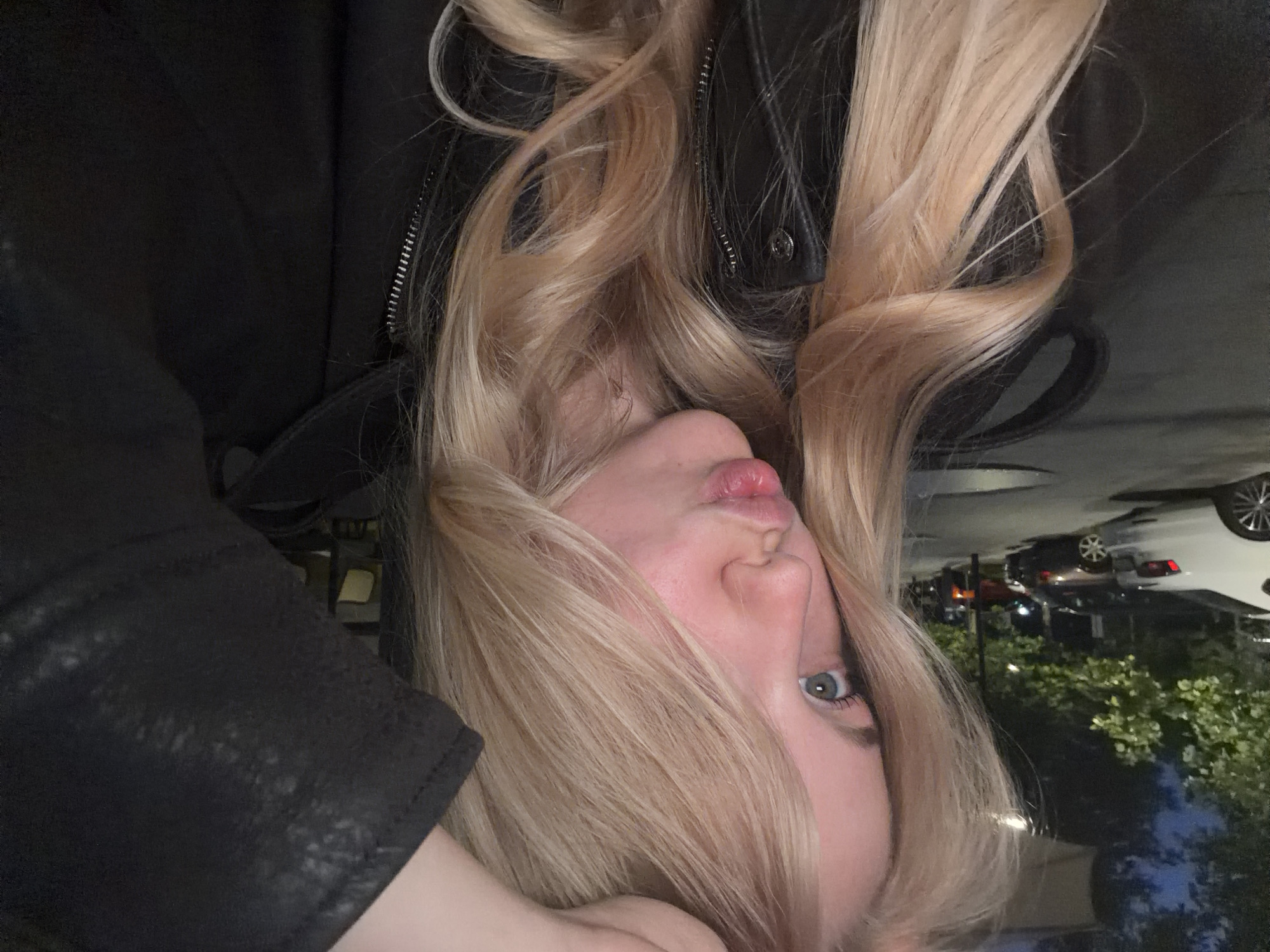 A woman with blonde hair and blue eyes is taking a selfie at night, possibly in an urban area, given the presence of parked cars and streetlights in the background. She is wearing a black jacket and has her hand on her head. She is puckering her lips as if to blow a kiss.