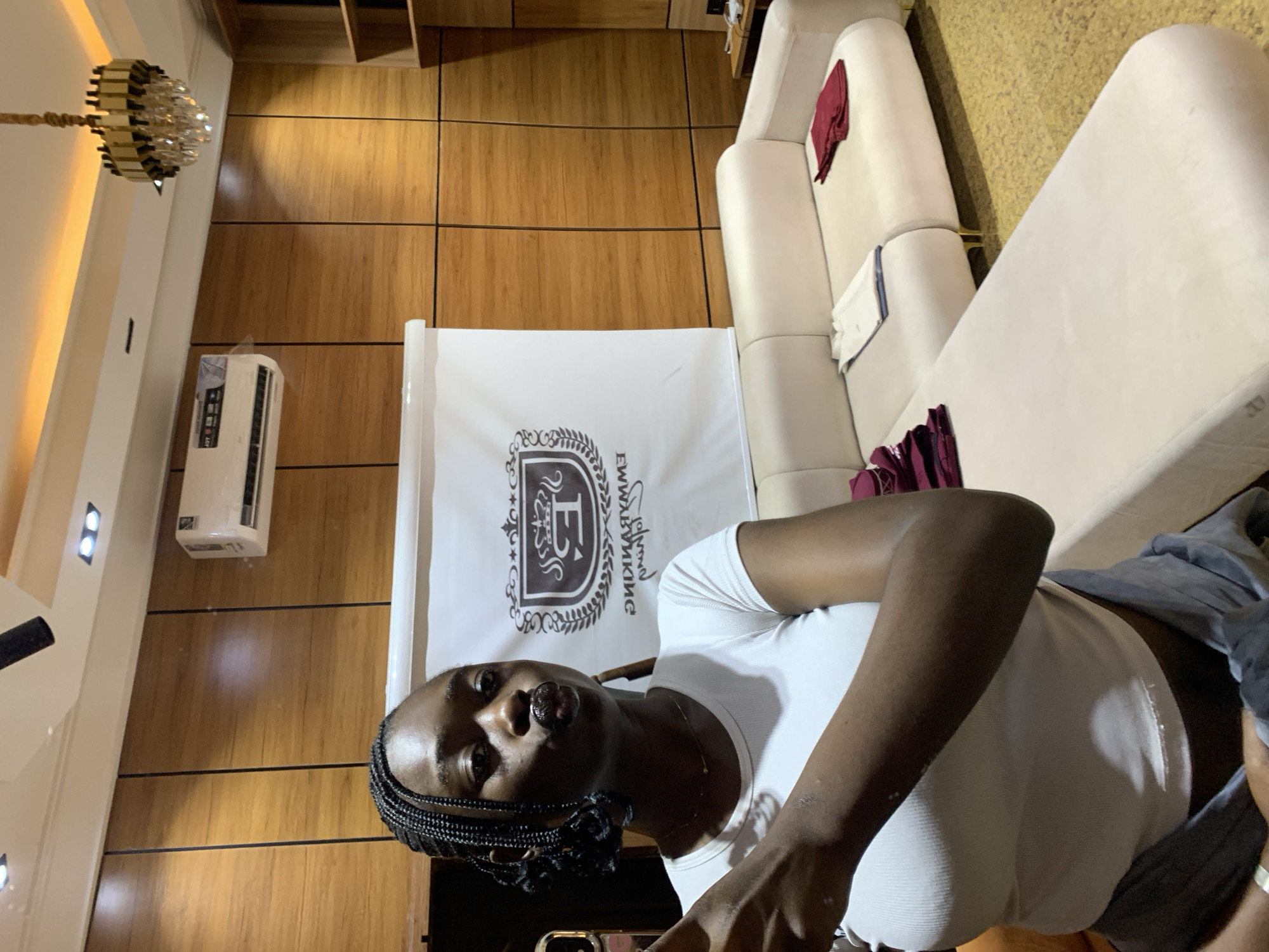 The image depicts an indoor scene featuring a young Black woman with braided hair, wearing a white crop top and grey pants. She is making a duckface pose while holding a mobile phone. Behind her, a white banner with a crest logo and text is displayed against a wood-paneled wall. To the right, a beige sectional sofa is visible, with folded burgundy and white garments placed on its cushions. Above the banner, a white air conditioning unit is mounted on the wall, and a modern chandelier hangs from the ceiling. The environment suggests a domestic or commercial setting, potentially a lounge area. The image does not provide any specific information about the city of Gura Top, Nigeria.