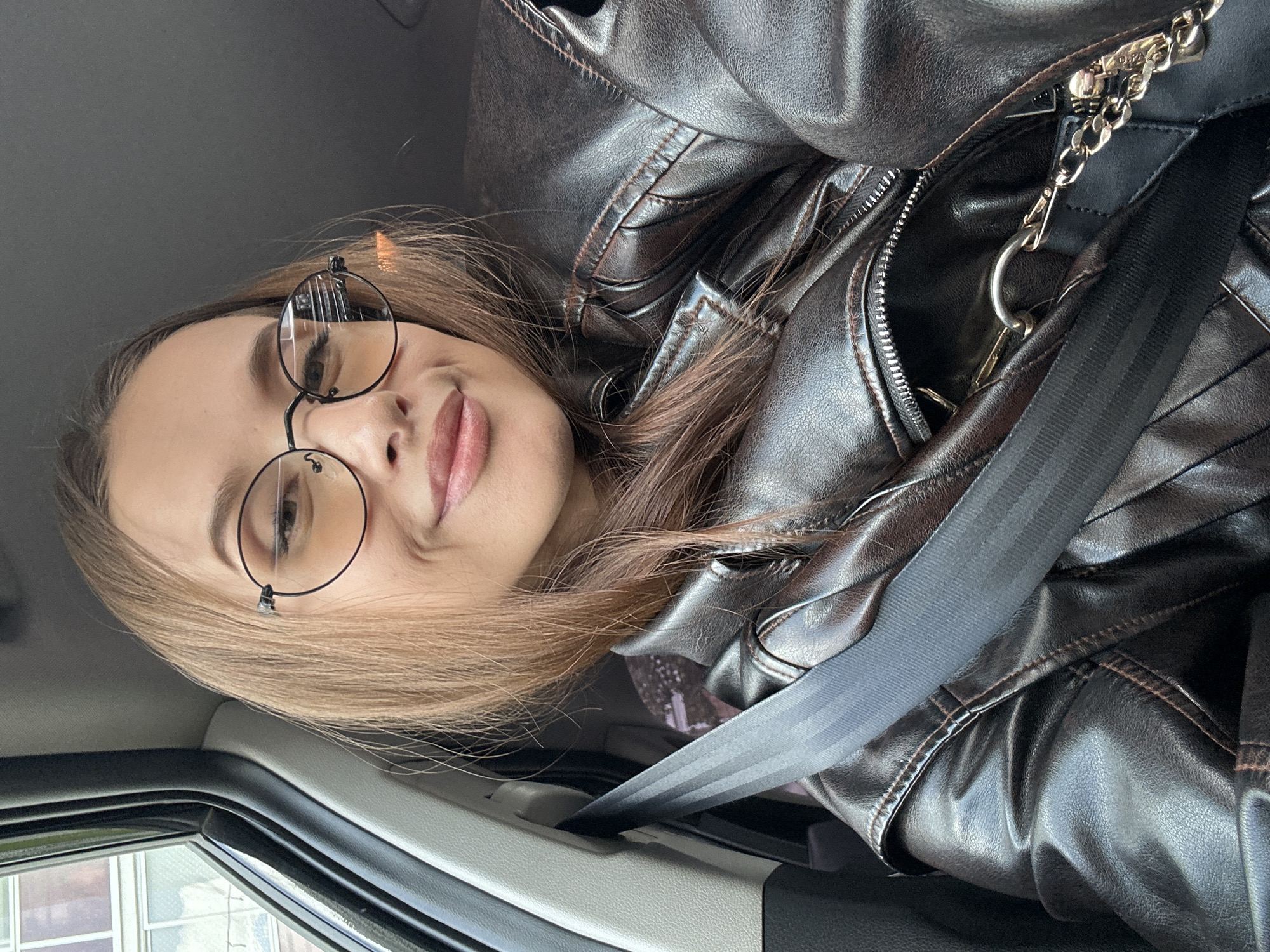 An adult female is depicted in a close-up, selfie-style photograph taken from inside a vehicle, specifically from what appears to be the front passenger seat. She has long, light brown hair, which is parted and falls over her shoulders. She is wearing round-framed glasses with a light amber tint and exhibits a slight smile while looking directly at the camera. Her lips are subtly made up, and her eyebrows are well-defined. She is dressed in a dark brown leather jacket with visible seams and a metallic zipper, partially open. A black seatbelt is securely fastened across her chest and lap. A black bag with a silver chain strap is visible beneath the seatbelt and jacket on her right side. The vehicle's interior features gray trim around the window and pillar on the left, and a dark gray headliner is visible above. Through the vehicle's window on the left, multi-story buildings with visible windows are partially visible, indicating an urban setting in the city of Yekaterinburg, Russia.