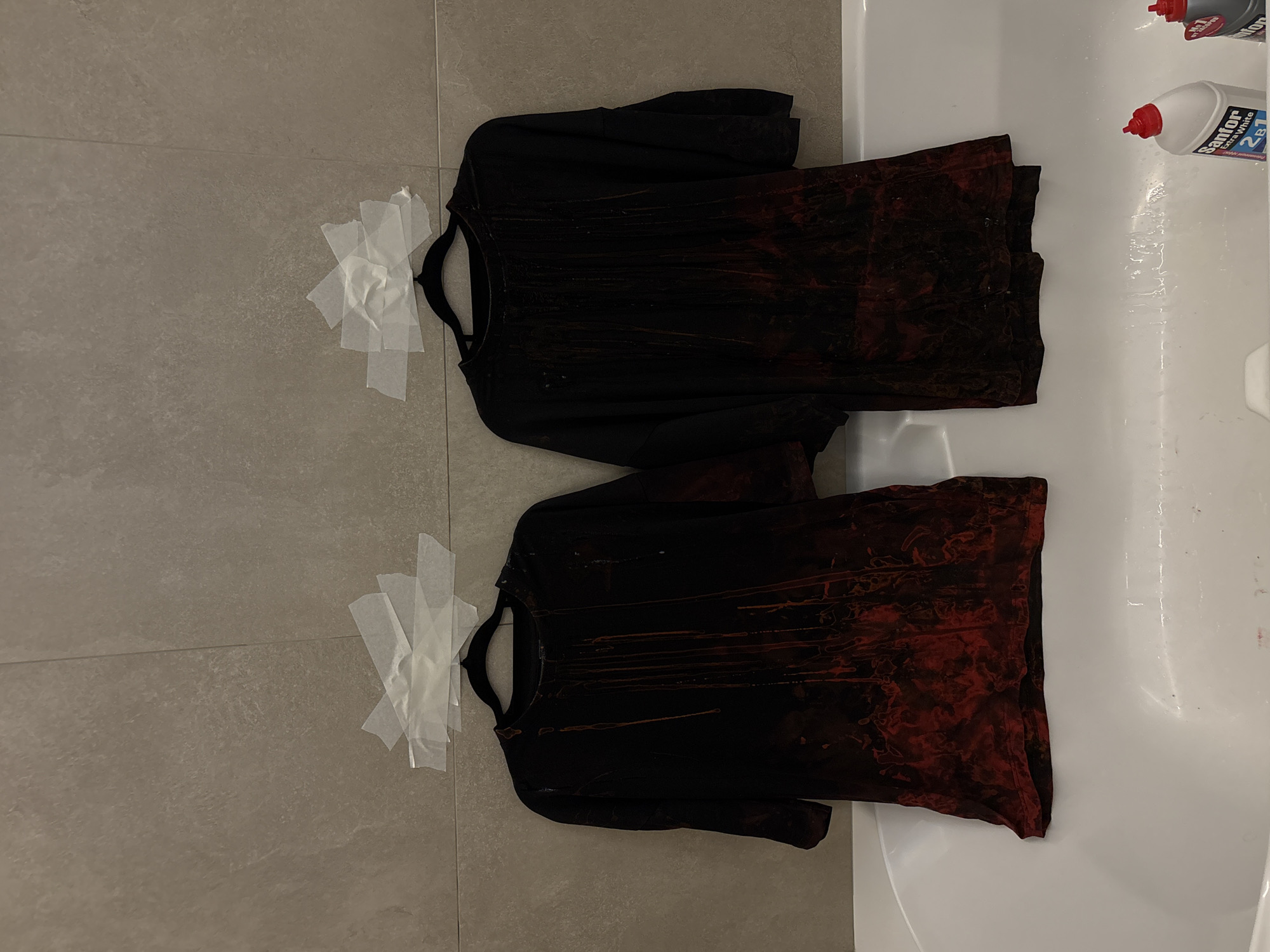 The image shows two black long-sleeved shirts splattered with red dye, hanging on hangers that are affixed to a grey tile wall using white tape. The shirts appear to be in a bathroom, as a portion of a white bathtub is visible to the right. Two bottles, one labeled "Sanfor Extra White," sit in the bathtub. The location is unknown as the scene shows a close up of a bathroom wall and tub with no landmarks visible.
