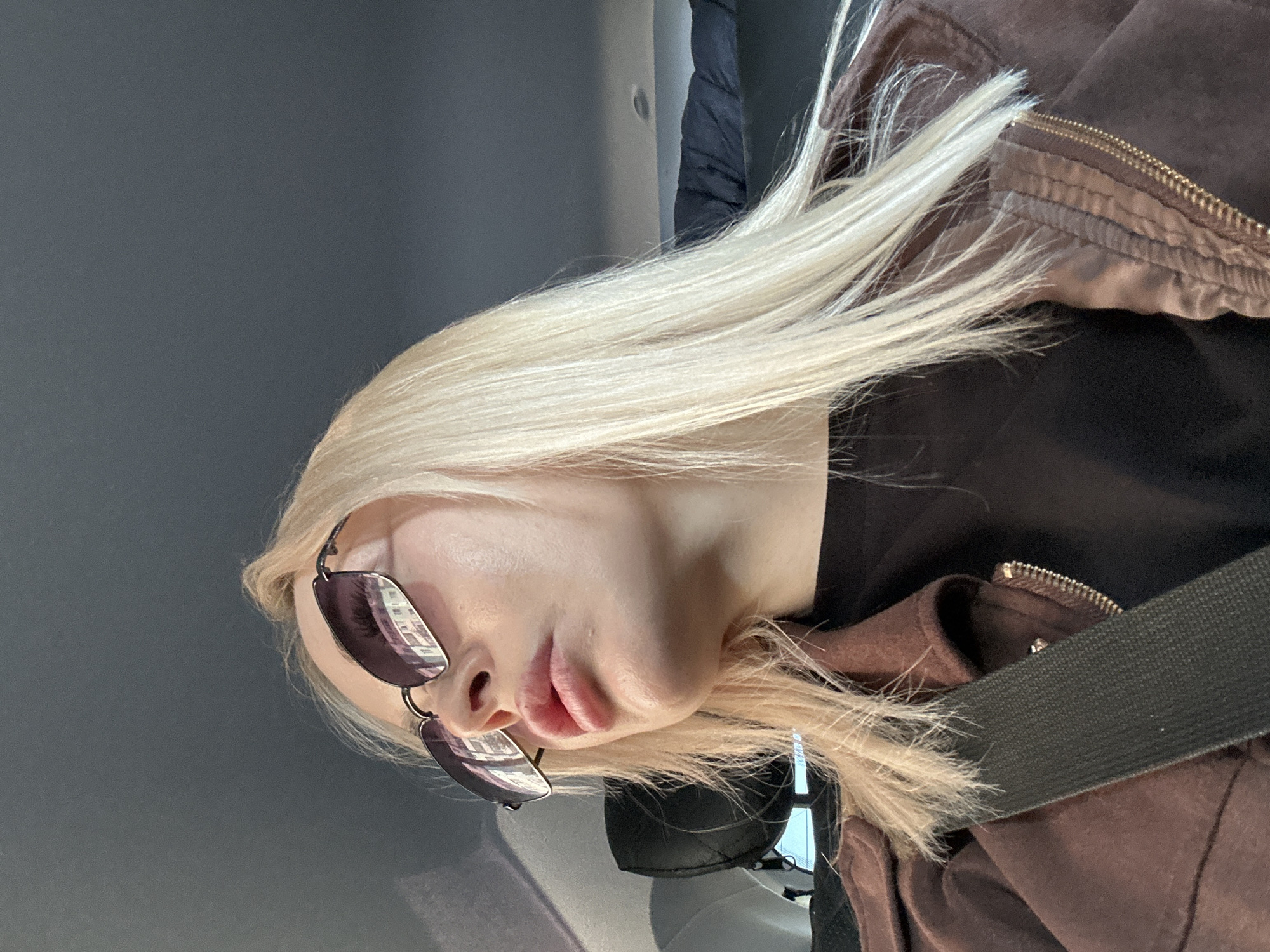 A blond-haired person wearing sunglasses and a brown jacket is seated in what appears to be a vehicle. A seatbelt is visible across their chest. Reflections of buildings are visible in the sunglasses. The background is a solid gray color, likely the interior of the vehicle. It is impossible to confirm the location as Megion, Russia from the given visual information.
