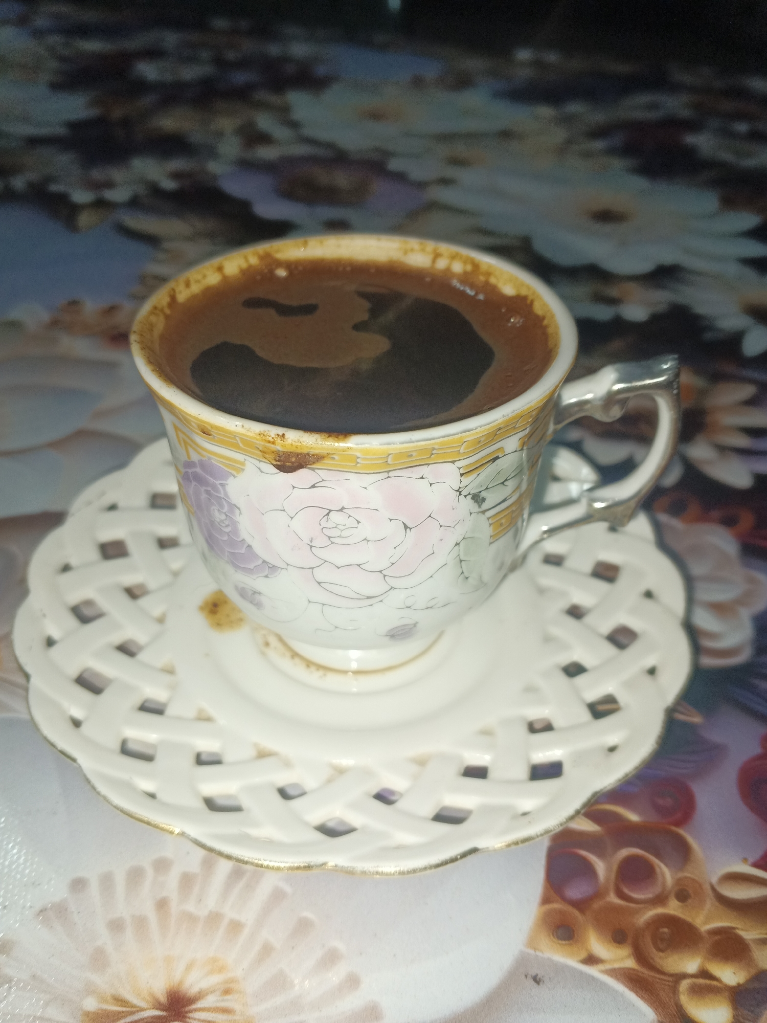 A close-up, overhead shot features a cup of coffee on a decorative saucer. The cup is white with a floral pattern of pink and purple flowers and gold trim. It contains dark coffee with foam. The saucer is white with an intricate, woven lattice design and gold trim around its edge. The background is a blurry, colorful surface with floral patterns. The overall scene appears to be indoors, possibly on a table. The image does not provide any information about the city of Sokhumi or Abkhazia.