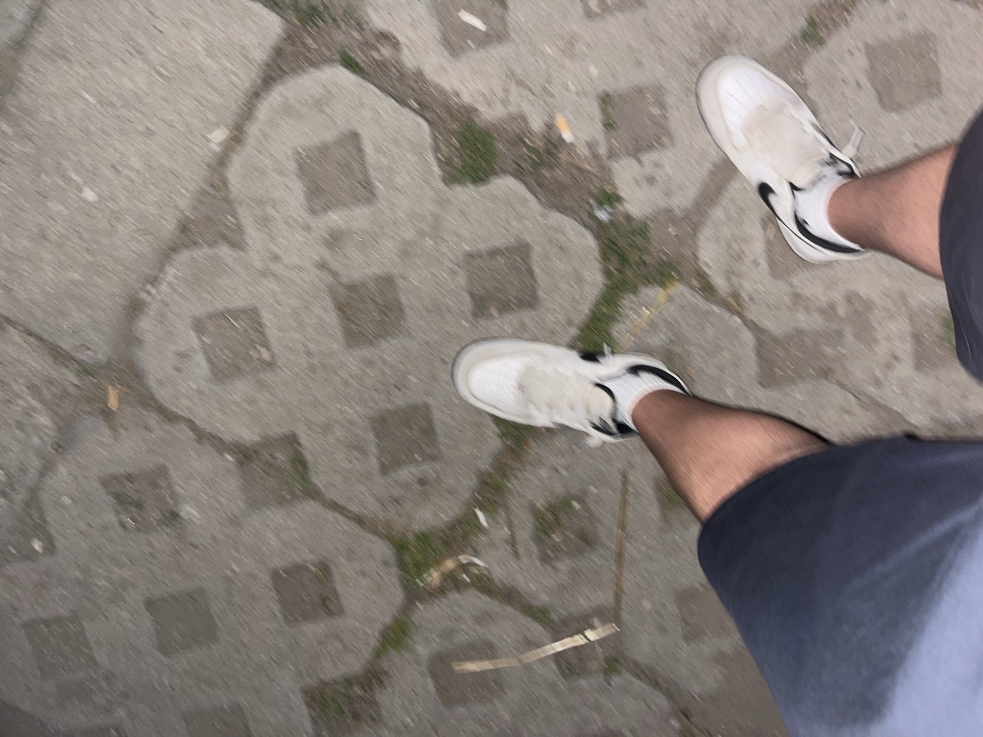 The image shows a pair of legs, wearing dark shorts and white sneakers, standing on a paved surface. The paving stones are irregular and have grass growing in the gaps between them. There are also some small pieces of debris scattered on the ground, including what appears to be a cigarette butt. The lighting suggests it might be evening or a dimly lit area. The scene is consistent with a pedestrian area in a park or a sidewalk in Plovdiv, Bulgaria.