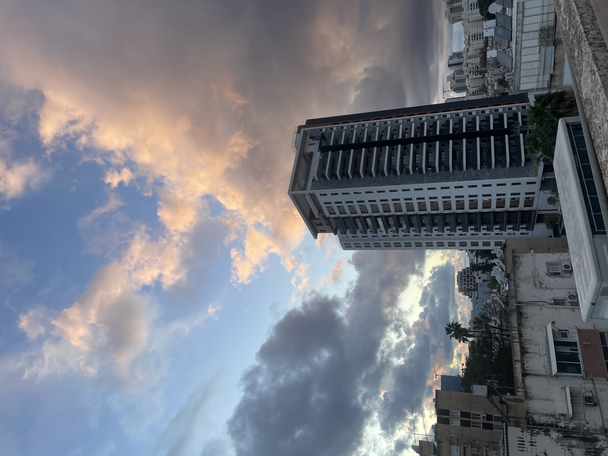 The image displays an urban landscape in Petah Tikva, Israel, captured from an elevated perspective, likely a rooftop or balcony. The scene is dominated by a dramatic sky featuring a mix of clear blue, dark grey storm clouds, and clouds illuminated with orange and pink hues, suggesting either sunrise or sunset.
In the mid-right, a tall, modern residential building stands prominently. It features a light grey facade, dark vertical accents, and numerous balconies with clear or frosted glass railings. The top level appears to have an open-air architectural design. To the left and extending into the midground, an array of older, multi-story buildings with varied architectural styles are visible. These buildings show signs of wear, with visible air conditioning units, exposed pipes, and rooftop water tanks and solar panels. A concrete ledge or wall, likely from the vantage point of the photographer, occupies the bottom portion of the frame, including a white, rectangular outdoor unit on its surface.
The urban environment includes sparse greenery, notably several palm trees, interspersed among the structures. In the distant background, additional buildings populate the horizon, and a construction crane is faintly visible, indicating ongoing development. No people, specific actions, or direct interactions are discernible within the frame. The overall lighting is characterized by strong contrasts between the brightly lit clouds and the shadowed cityscape.
