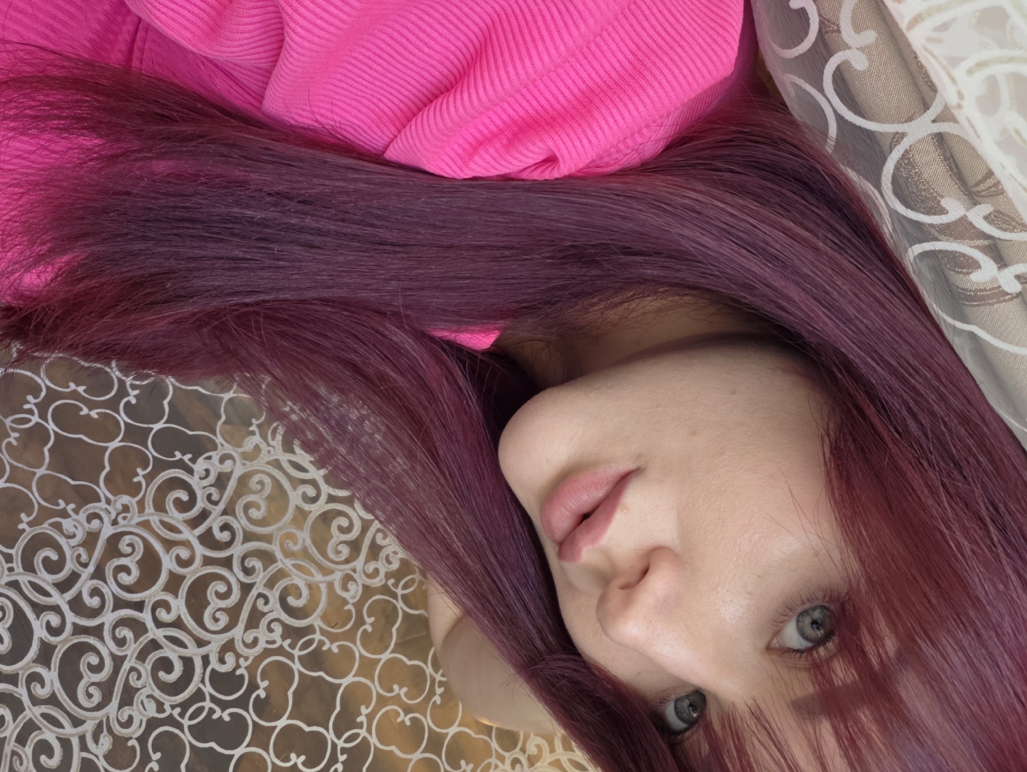 A close-up, eye-level, medium shot features a young Caucasian woman with long, dark plum colored hair and a fringe. She is looking directly at the camera, with her head tilted to her left. Her eyes are a pale grey-green, and her lips are painted a soft pink. She is wearing a bright pink ribbed hooded sweatshirt. Behind her, to the right, a patterned fabric with white swirls on a gold background is visible. To the left of the frame, a beige and white patterned sheer curtain is partially visible. The image appears to be taken indoors. There is no specific identifiable location in the city of Chaikovsky, Russia.