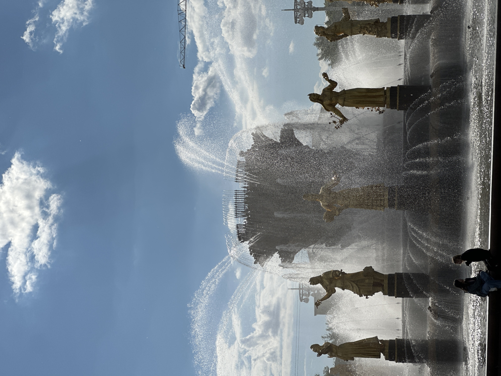 The image captures a vibrant, sunny scene at a grand outdoor fountain, likely the "Friendship of Nations Fountain" at VDNKh in Moscow, Russia. The fountain dominates the frame, featuring multiple golden statues of female figures, each standing on an ornate pedestal. These statues are integral to the fountain's design, which unleashes powerful jets of water that create a spectacular, glittering spray across the scene. The water catches the sunlight, creating bright reflections and a misty atmosphere around the statues.
In the lower right portion of the frame, two individuals are visible, silhouetted against the bright spray of the fountain. One person appears to be bending or leaning forward, while the other is in a sitting or kneeling posture, seemingly observing or interacting with the water.
The background consists of a clear blue sky, adorned with scattered white cumulus clouds. A tall, dark metal structure, possibly part of a crane or an adjacent monument, is visible against the sky in the upper left. The bright daytime sun casts strong highlights on the water and statues, creating a dynamic interplay of light and shadow throughout the scene. There is no visible text in the image.