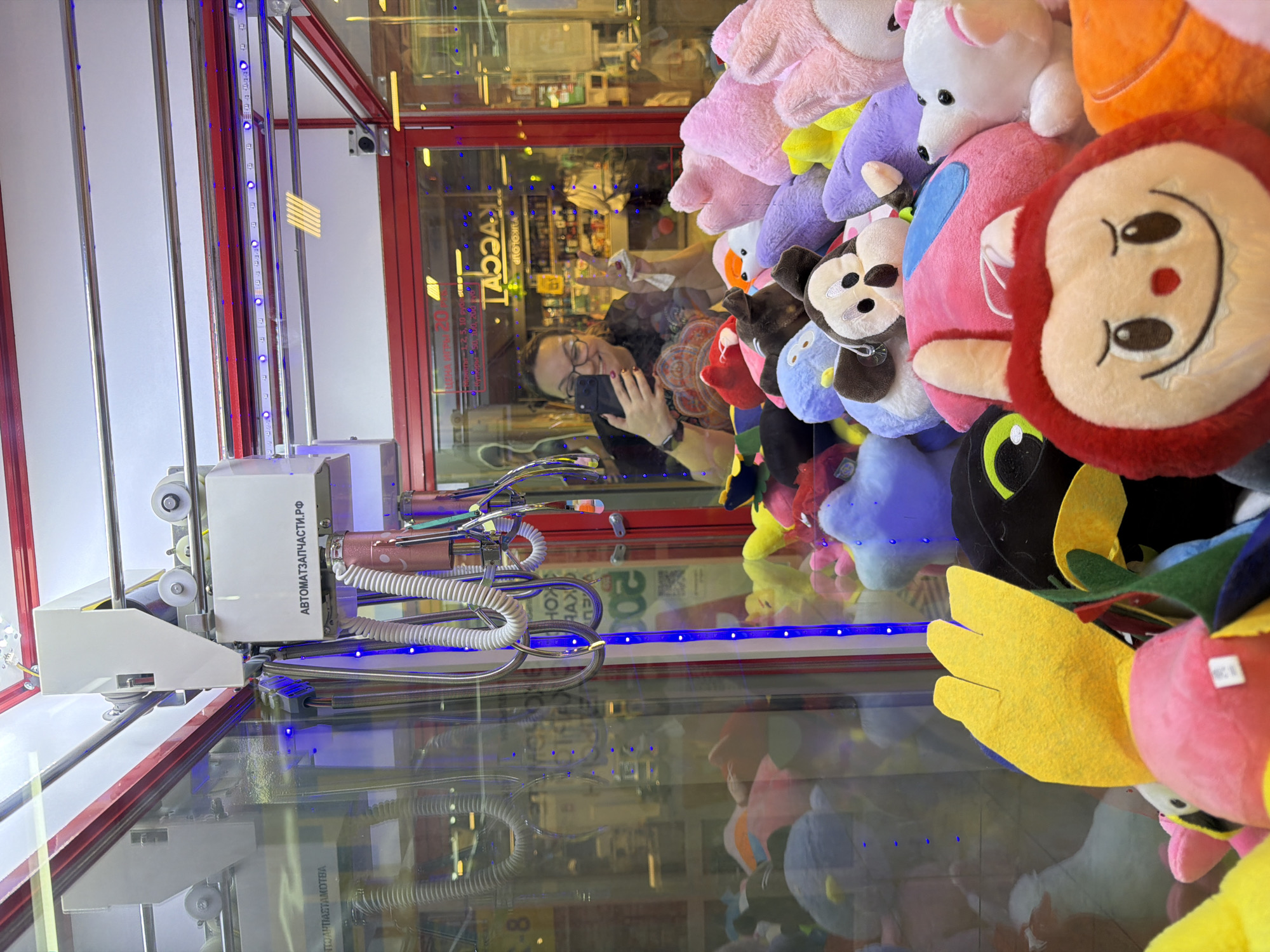 Here is a detailed description of the image:
**Scene:** An indoor environment, likely an arcade or entertainment center, in the city of Chaikovsky, Russia. The focus is on a claw machine filled with plush toys.
**People:** A woman is reflected in the glass of the claw machine. She is wearing glasses, a patterned shirt, and a watch. She is holding a smartphone and appears to be taking a picture of herself reflected in the glass of the machine. She is also making a peace sign with her fingers.
**Subjects:** A variety of colorful plush toys are visible inside the claw machine. Some are distinct characters, while others are generic animal shapes.
**Objects:**
* The claw machine itself, with its metal frame, glass enclosure, and mechanical claw mechanism. There are blue LED lights around the inside of the machine.
* A control unit with writing in Russian on it.
* The smartphone being held by the woman.
**Actions:** The woman is taking a selfie in the reflection of the claw machine.
**Interactions:** The primary interaction is between the woman and her phone, as well as her interaction with the reflection of herself in the claw machine.
**Environment:** The setting is an arcade, which is implied through the presence of the claw machine. The reflections in the glass suggest the arcade is somewhat busy, with other lights and objects in the background.