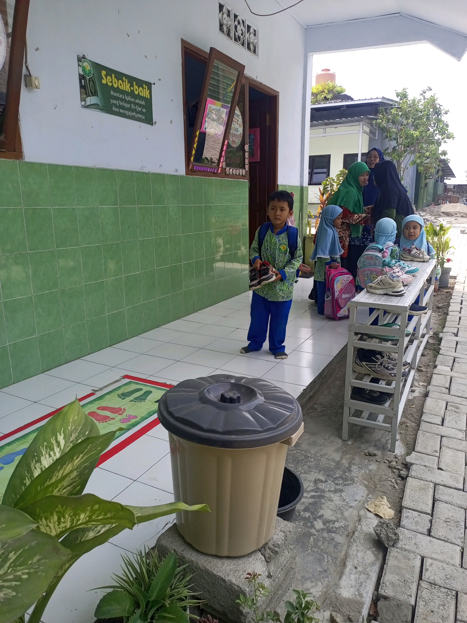 The image depicts an outdoor scene in front of a building, likely a school, in Gurah, Indonesia. In the foreground, a young boy in a blue and green uniform stands holding a pair of black and red shoes. To his right, a white shoe rack displays several pairs of shoes, and beyond that, a group of people, including women in hijabs and children, are gathered. One woman, wearing a green hijab, is interacting with the others. The building features a green tiled wall and a white overhang. A large beige trash can is visible on the left side of the image, next to a green rug with footprints. The ground is a mix of tiled and paved surfaces. The overall environment appears to be an urban or semi-urban setting.