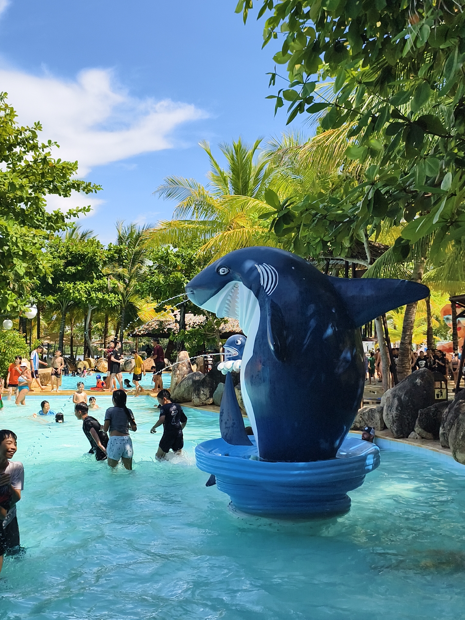 The photograph captures a vibrant scene at a water park located in Tay Nha Trang Ward, Vietnam. The foreground prominently features a large, blue and white shark statue mounted on a tiered, blue base within the water. Children of varying ages are seen playing in the pool. Some are wading through the water, while others are swimming. The background is filled with additional park goers and lush tropical vegetation, including palm trees and leafy trees, under a partly cloudy blue sky. Large rocks line the edge of the pool.