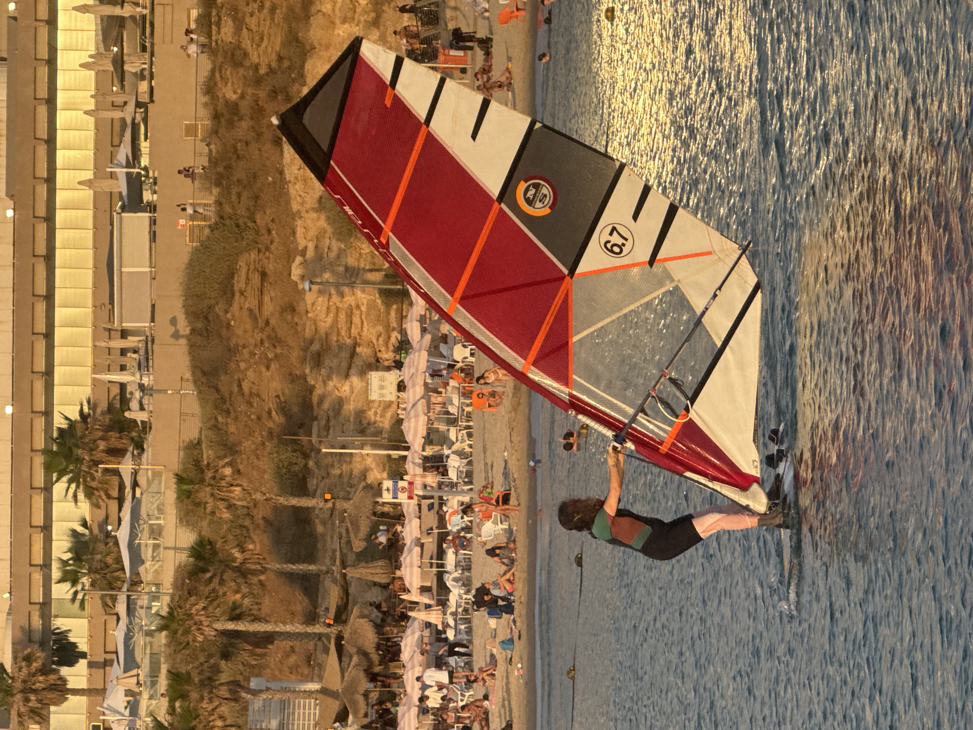This image depicts a vibrant late afternoon scene at a beach in Tel-Aviv, Israel, characterized by a distinct sandy cliff leading to the shoreline.
In the immediate foreground, a person is windsurfing on calm, ripple-patterned water that reflects the golden light of the setting sun. The windsurfer, wearing a dark short wetsuit with a green and orange panel on the shoulder, stands on a white board. They are holding the boom of a large windsurf sail. The sail is predominantly red, white, and black, with orange batten pockets and features a circular logo containing "N S" and the number "6.7" prominently displayed. Other individuals are visible in the water, some swimming or wading.
The midground reveals a crowded sandy beach lined with numerous white beach chairs and various umbrellas. These include many striped umbrellas, primarily pink and white, and several natural straw-thatched umbrellas. A multitude of people are scattered across the beach, relaxing, walking, and engaging in various leisure activities. A light pole and a warning sign (featuring a red circle with a diagonal line) are visible near the back of the beach area.
The background is dominated by a sandy cliff or hill, heavily vegetated with numerous palm trees. Above the cliff, an elevated promenade or walkway extends horizontally, with several individuals visible on it, some leaning against railings. Further back, a large multi-story building, likely a hotel or residential complex, stands prominent. Its facade, possibly made of glass or reflective material, is illuminated by the golden sunlight, giving it a warm, metallic sheen. Artificial lights are visible along the building and the promenade. The overall lighting suggests the golden hour, with warm, directional light casting soft shadows and creating shimmering reflections on the water.