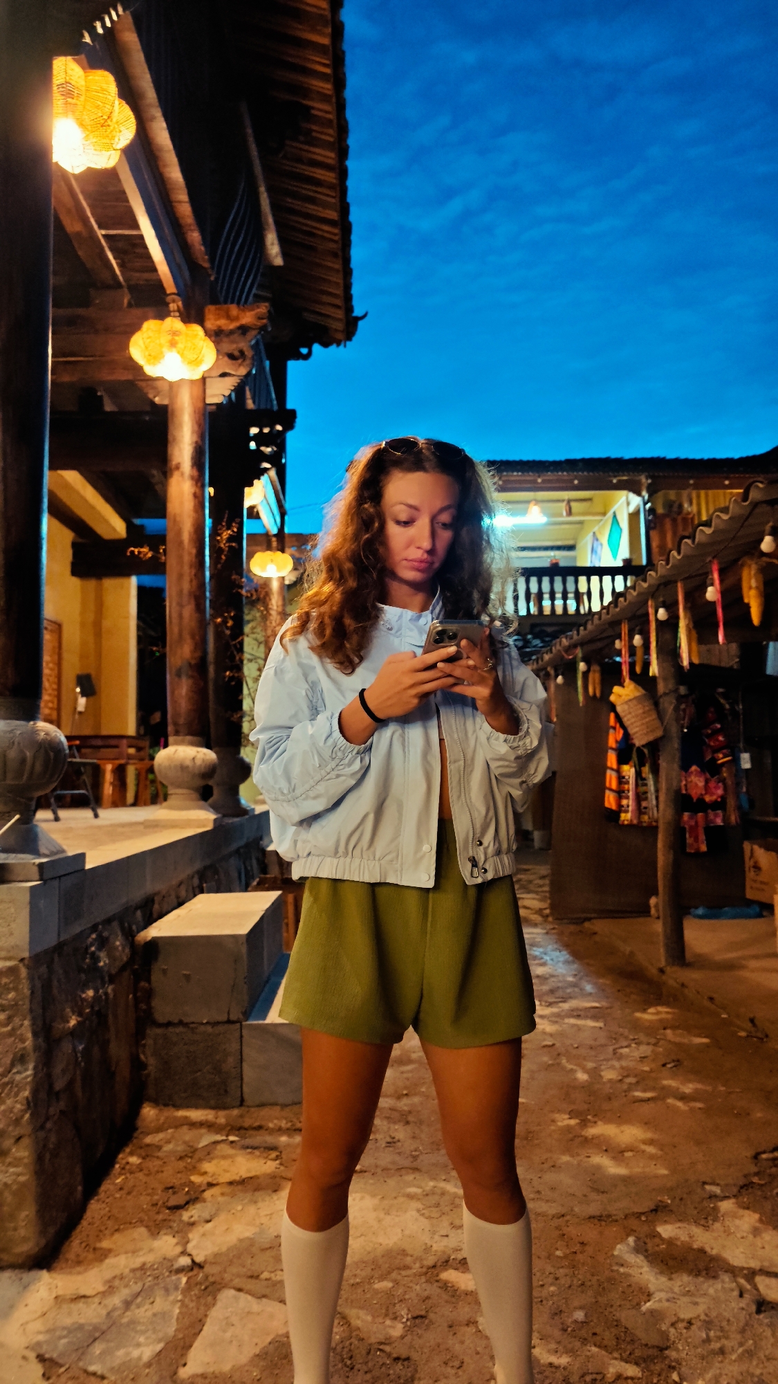 A woman with curly brown hair stands on a stone walkway, looking at her phone, in Lũng Cú Commune, Vietnam. She is wearing an unbuttoned light blue jacket, olive green shorts, and white knee-high socks. The lighting is dim, with yellow lanterns illuminating the area. The background includes traditional Vietnamese architecture with wooden structures and hanging decorations. The sky is a deep blue, indicating it is dusk or evening.