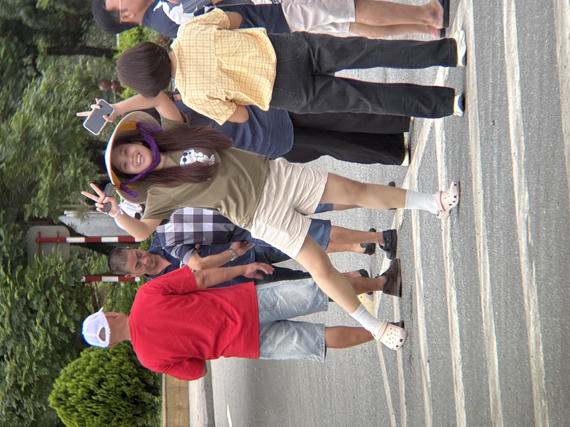In Lao Bảo Commune, Vietnam, a young woman in a conical hat and a green t-shirt is captured in a dynamic pose, holding a smartphone and making a peace sign with her left hand. She is wearing light-colored shorts and pink slip-on shoes. Behind her, a man in a red t-shirt and denim shorts stands facing away from the camera. To his right, another man in a checkered shirt is partially visible. Further right, a woman in a yellow checkered shirt and black pants is facing away from the viewer. The background consists of trees and a street scene with pedestrian crossing lines on the asphalt. The overall atmosphere suggests a casual outdoor gathering or stroll within the town.