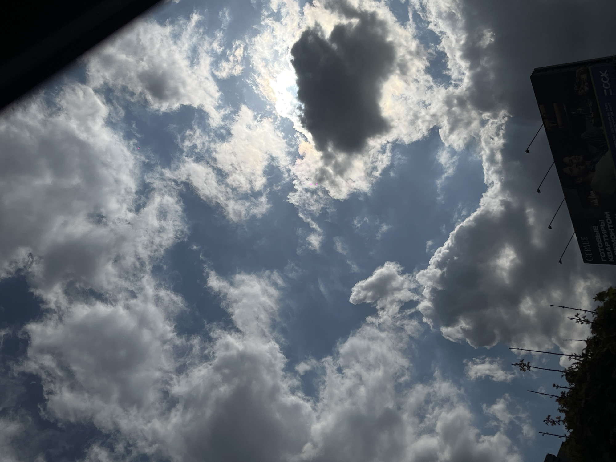 The image presents an upward-looking view of a dynamic, partly cloudy sky, characteristic of a daytime scene. The sun, partially obscured by dense clouds in the upper-center, casts bright light and sunbeams through gaps, creating a striking contrast between illuminated and shadowed cloud formations. A large, dark, somewhat heart-shaped cloud dominates the central sky, blocking much of the direct sunlight. The clouds vary in appearance, from wispy white to heavy, dark grey cumulus, suggesting a changeable weather pattern.
No specific human activity is visible in the sky. In the upper-left corner, a dark, diagonal element, likely part of a car's interior or another structure, cuts across the frame. On the far right side, a tall billboard is partially visible, oriented vertically and mostly in shadow, with text printed in Cyrillic. Below the billboard, a dark silhouette of tree branches or foliage is noticeable in the bottom-right corner.
Notable details include the dramatic interplay of light and shadow created by the sun and clouds. The visible text on the billboard provides significant location cues: "СБС," "ГКК," "ГОТОВЫЕ КВАРТИРЫ" (Ready apartments), "СЕРИЯ," "КРАСНОДАР," and "РЕМОНТОМ" (with renovation). The mention of "Krasnodar" confirms the provided location context. The billboard is advertising real estate. The time of day appears to be mid-day due to the high position and intensity of the sun, despite its partial concealment.