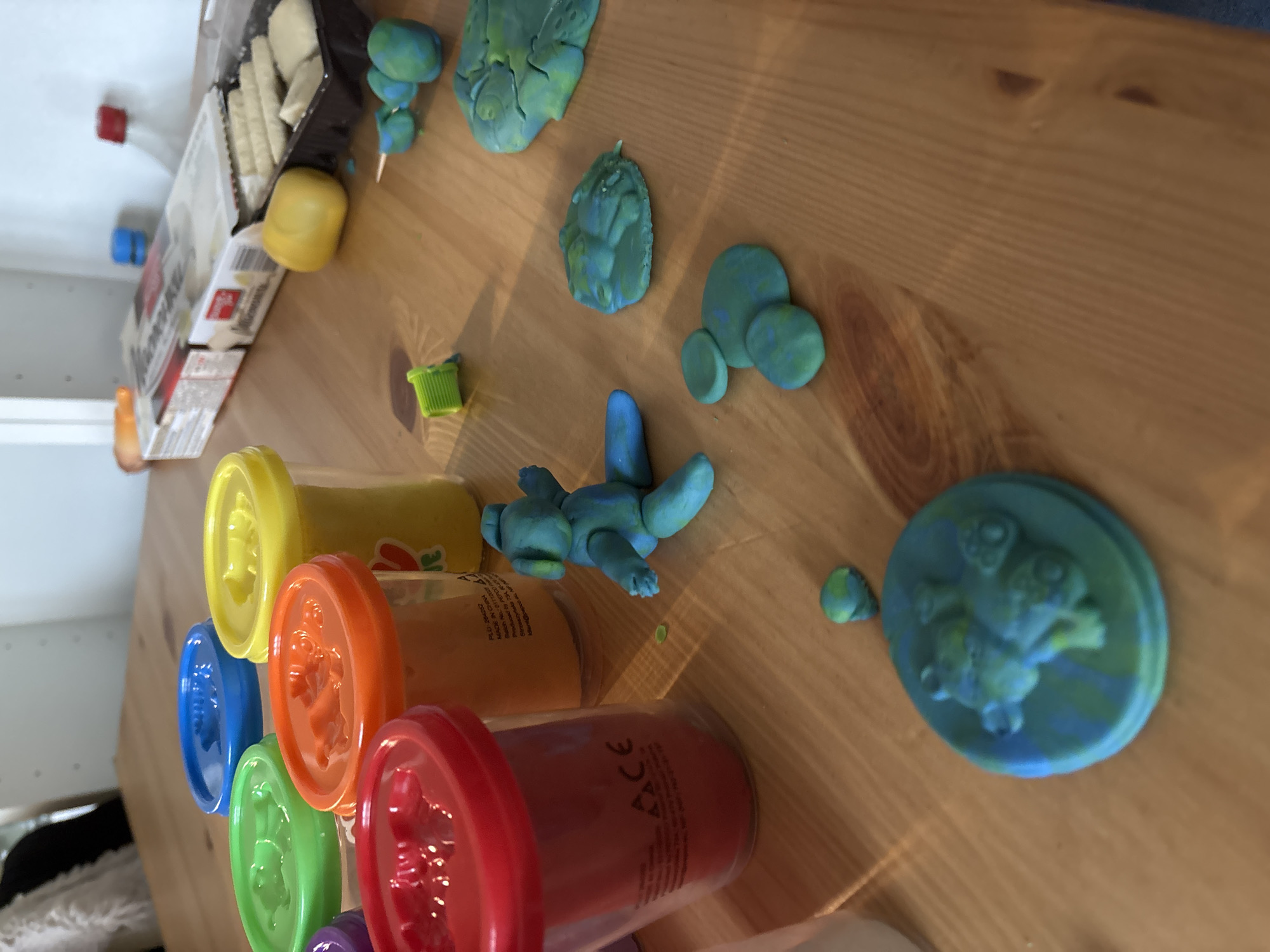 On a light brown wooden surface, a variety of objects are arranged. The left side of the frame features stacked containers of colorful play dough in yellow, orange, red, blue, green and purple. Next to these are items appearing to be play dough creations in shades of blue and green. A box of wafer biscuits and a yellow plastic container are near the top left of the frame. The lighting in the image is moderate and the focus is sharp, allowing for clear detail of the items present. There is no evidence to suggest that the image was taken in Sofia, Bulgaria.
