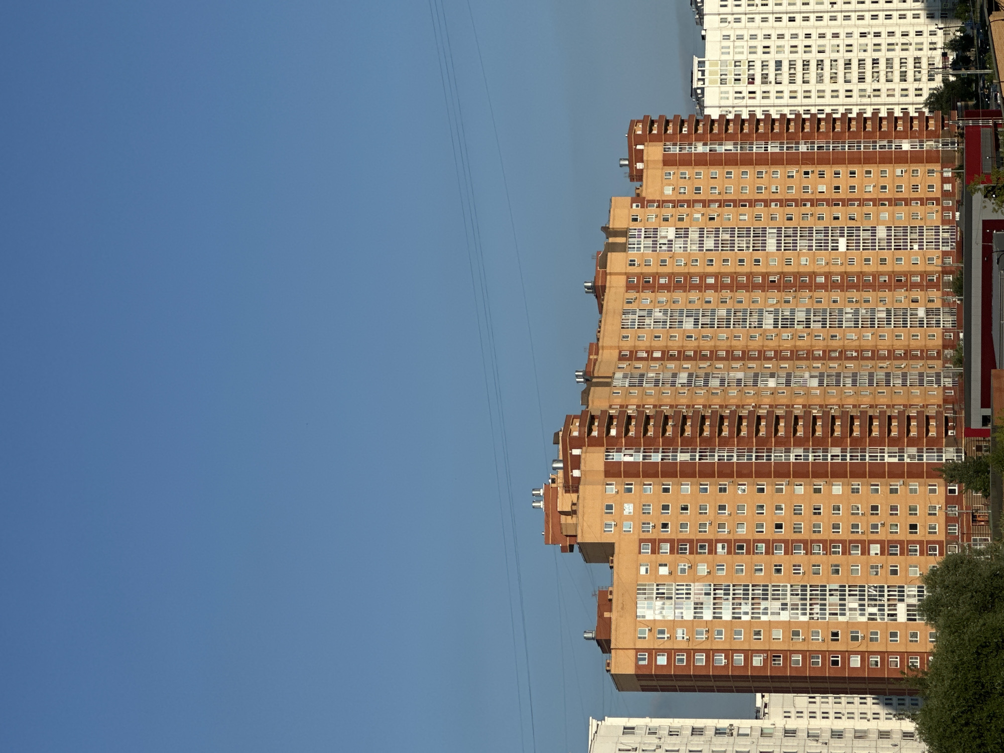 The image shows a section of the city of Podolsk, Russia, with a clear blue sky dominating the upper half of the frame. Below the sky are several tall apartment buildings. The most prominent building is a large, multi-story residential complex with a distinctive orange and reddish-brown facade. This building features numerous windows, balconies, and what appear to be air conditioning units mounted on the exterior. To the left of this building is another, taller, white apartment building. To the right, a white, multi-story building is partially visible, also with many windows. Thin power lines can be seen stretched across the sky above the buildings. In the foreground, a line of green trees obscures the lower portion of the buildings. The overall scene depicts a modern urban residential area under a clear sky.