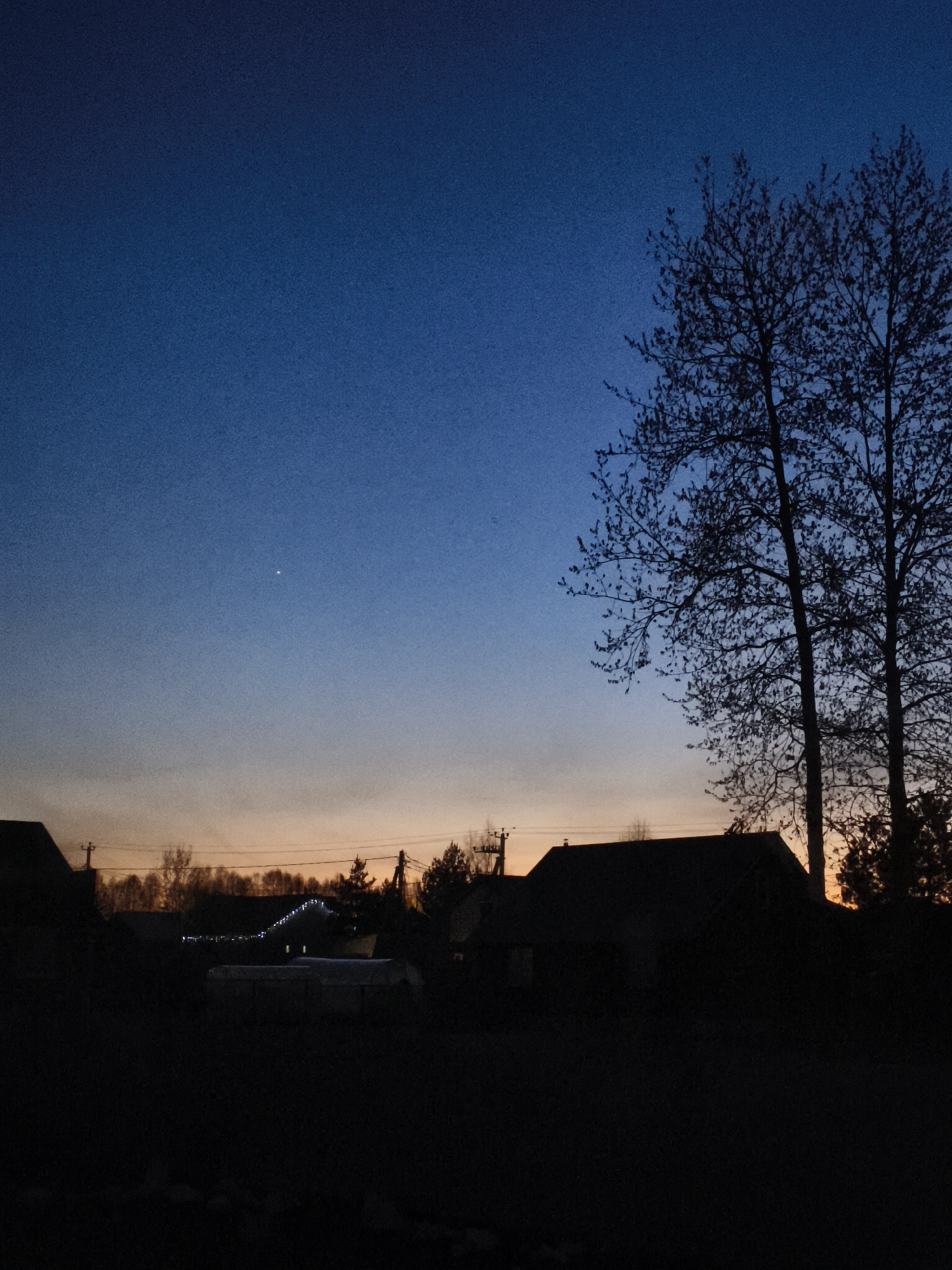 The image depicts a twilight or early evening scene in Русский Вожой, Russia. The sky transitions from a deep blue at the top to an orange-tinted horizon. A single bright star or planet is visible in the upper left portion of the sky. Silhouetted trees are on the right side of the image. Below the horizon, silhouettes of houses and other structures are visible, as well as power lines. The foreground is dark, suggesting a field or yard. There are lights on the top of a house.