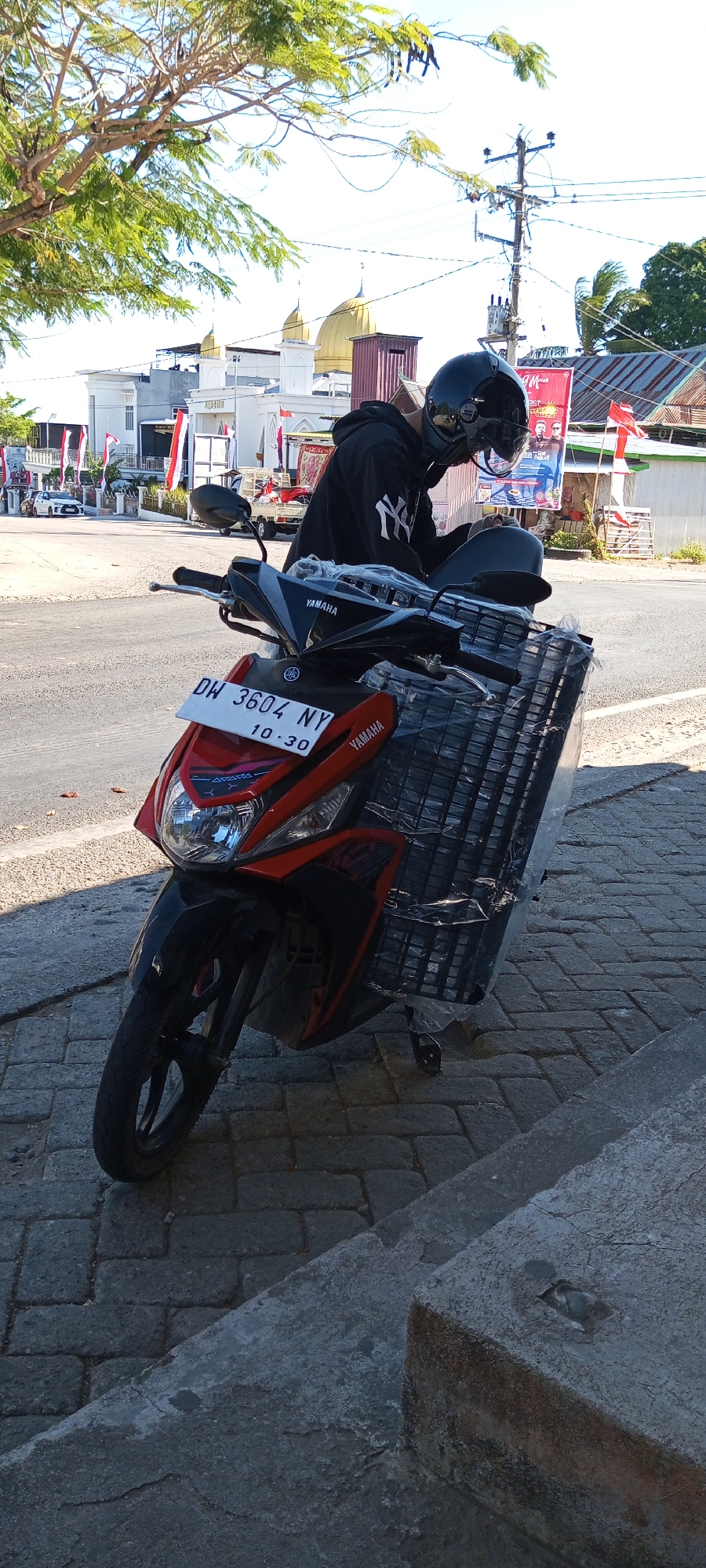 An exterior medium shot captures a red Yamaha scooter parked on a paved sidewalk adjacent to a road. A person wearing a black hooded sweatshirt and a black helmet is seated on the scooter, their head bowed. The scooter's front displays a license plate that reads "DW 3604 NY 10.30". A large plastic-wrapped basket is attached to the rear of the scooter. In the background, buildings with white facades and red-striped flags are visible, along with a golden-domed structure and a utility pole with electrical wires. The scene is illuminated by bright daylight, suggesting it is daytime.