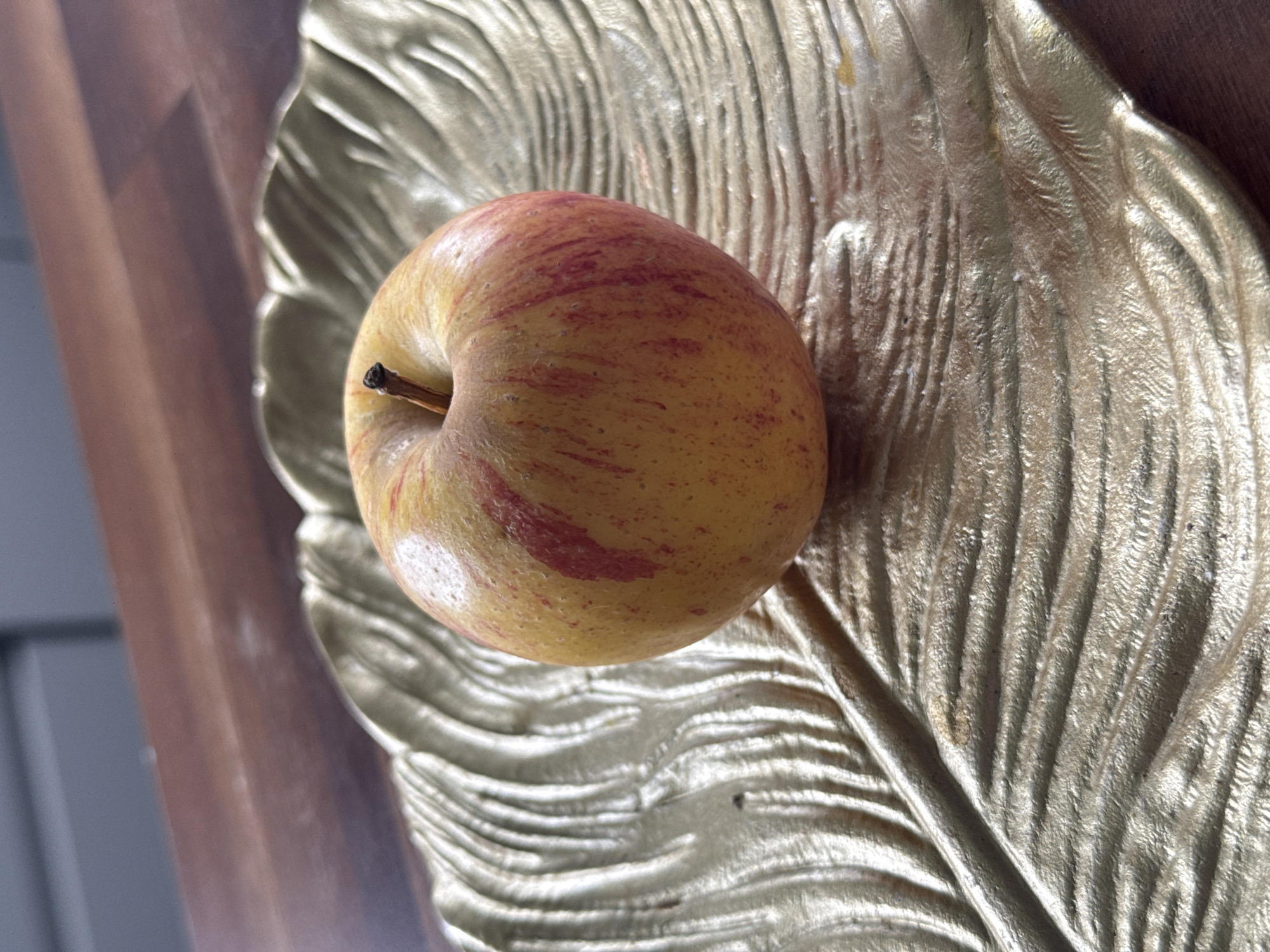 An apple rests on a golden, leaf-shaped dish. The dish is textured with raised lines that mimic the veins of a leaf. The apple is mostly yellow with streaks of red. The dish is placed on a wooden surface. The background is blurred, showing what appears to be a kitchen counter and cabinetry. The scene is indoors, likely in a home. The location in Птицевод-2 Снт, Russia is not discernible from the image.