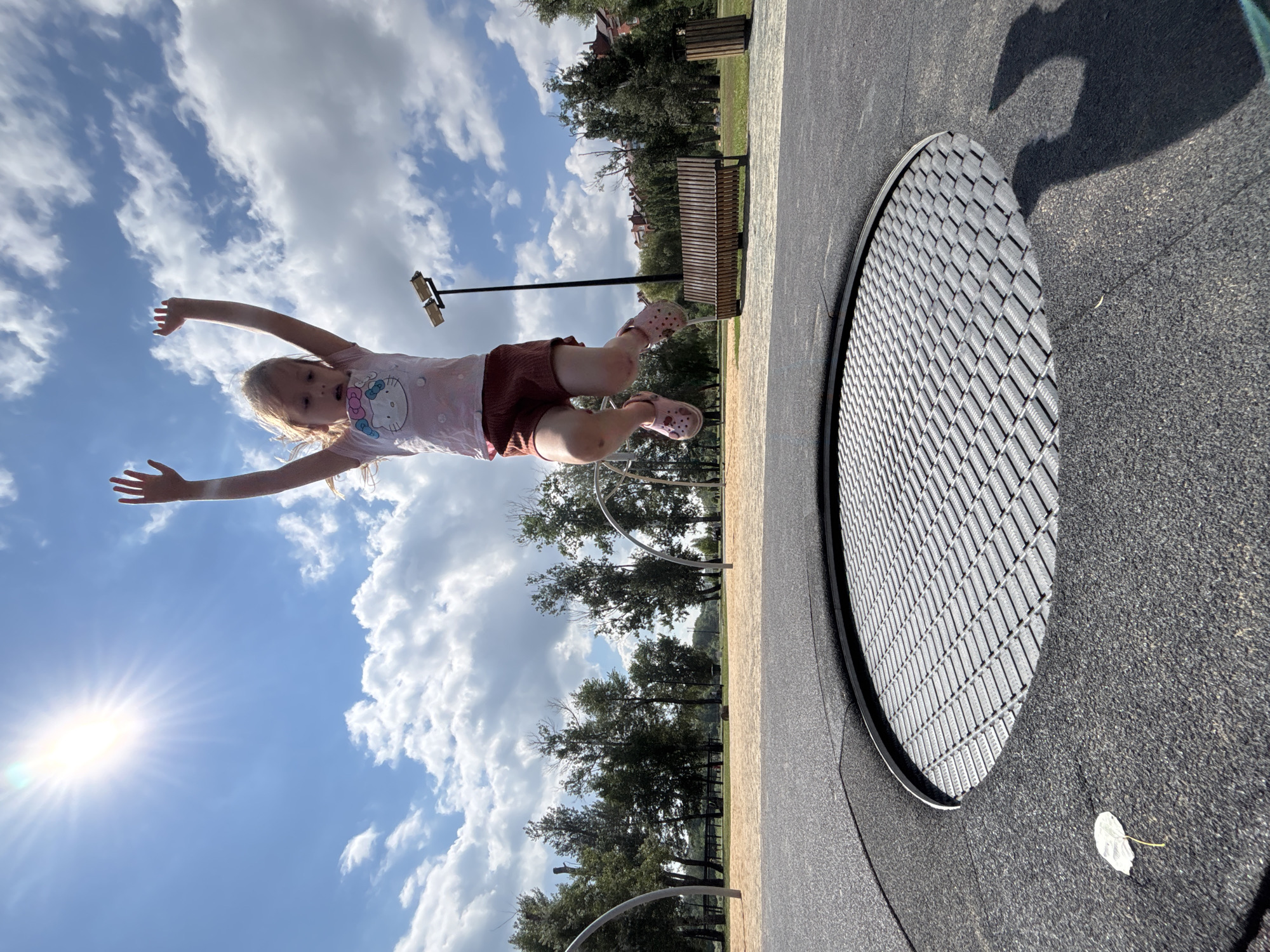 An outdoor, ground-level trampoline is in the foreground of an image. A young girl with blonde hair is captured mid-air, jumping on the trampoline. She is wearing a light pink t-shirt with a cartoon character and dark red shorts, along with pink clogs. Her arms are raised above her head. The trampoline is circular with a metal mesh surface and a black rubberized border. The background shows a park area with sandy ground, trees, a bench, and a lamppost. The sky is blue with scattered white clouds, and the sun is shining brightly in the upper left corner. The image was taken in the city of Orenburg, Russia.