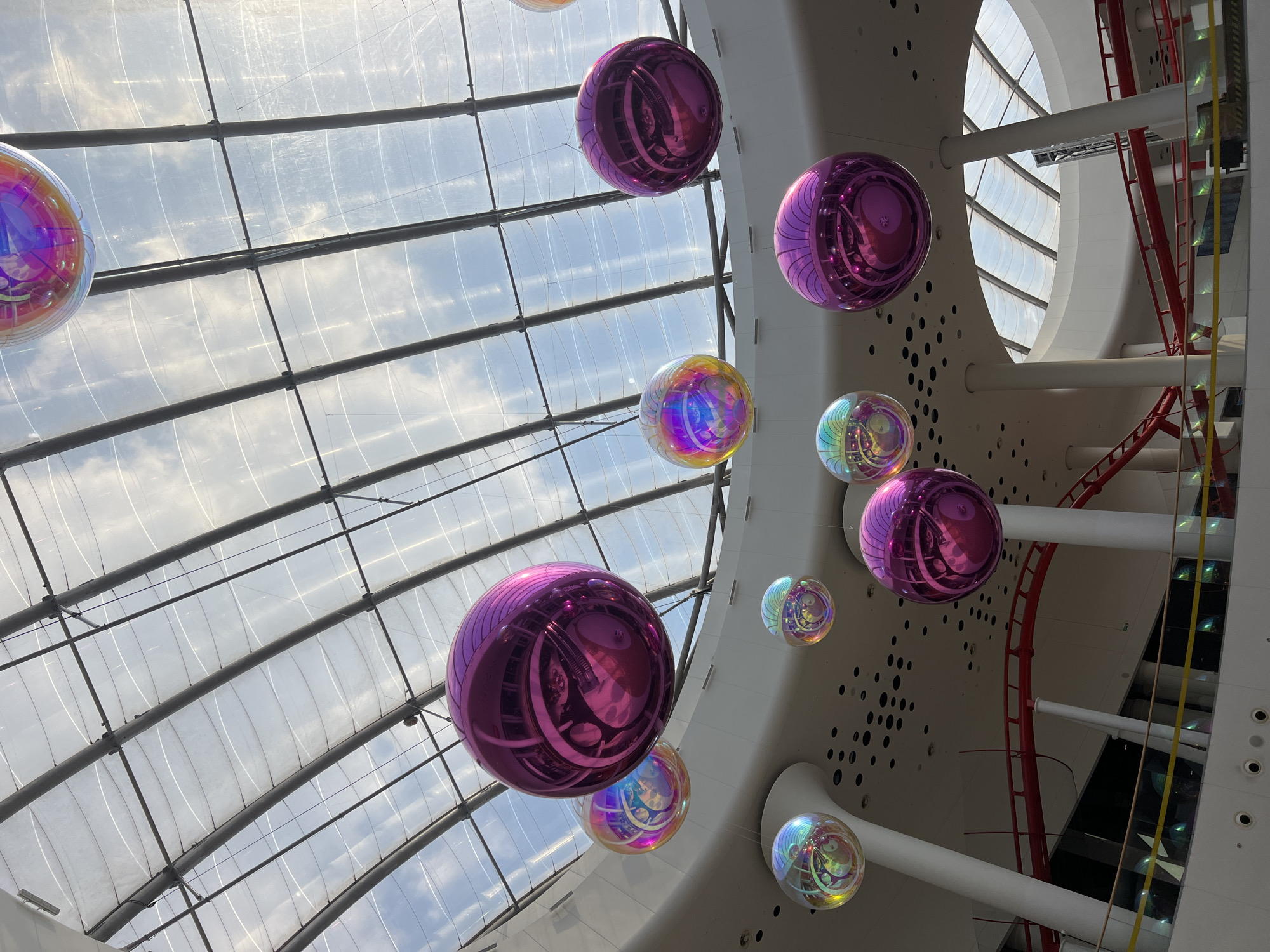 The image shows a large, multi-level interior space with a translucent, curved roof. Hanging from the ceiling are several large, reflective, spherical decorations. Some are a bright, metallic purple, while others are iridescent and show a rainbow of colors. The reflections on the spheres indicate an indoor environment with artificial lighting and possibly some natural light filtering through the roof. Below the spheres, architectural elements are visible, including white columns, curved balconies with railings, and a red roller coaster track. The overall impression is of an entertainment venue or a shopping mall.
The prompt requests the location be in Kyiv, Ukraine, however, based solely on the visual information present in the image, it is not possible to determine the specific location within Kyiv, Ukraine or any other city. There are no identifying landmarks or signs to confirm the location.