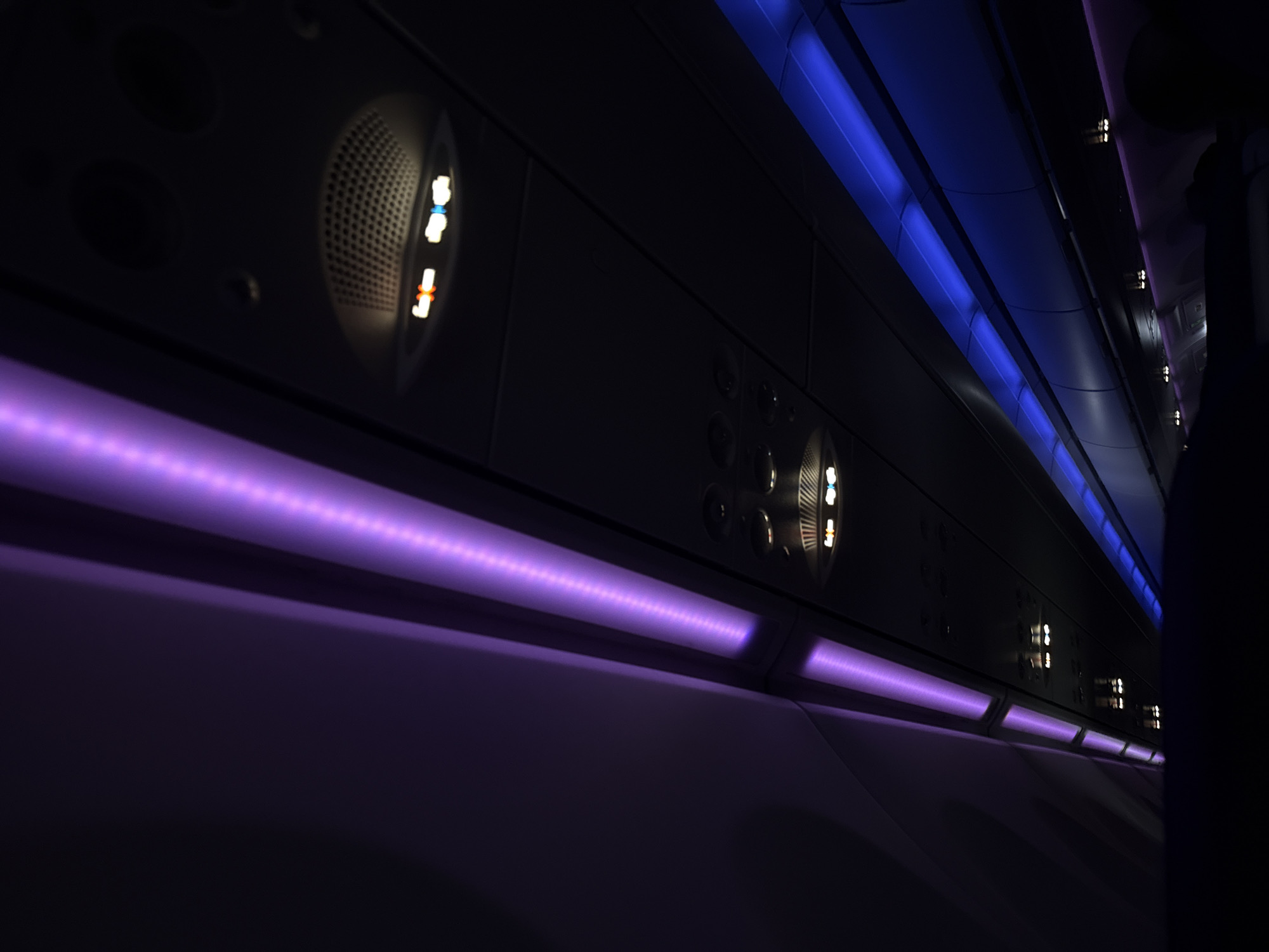 The image depicts the interior of an airplane cabin at night, illuminated with purple and blue LED lighting. Overhead panels display individual air vents and passenger controls (call button and reading light). The scene is dark, and details are obscured by shadows. The image is not linked to Yerevan, Armenia.