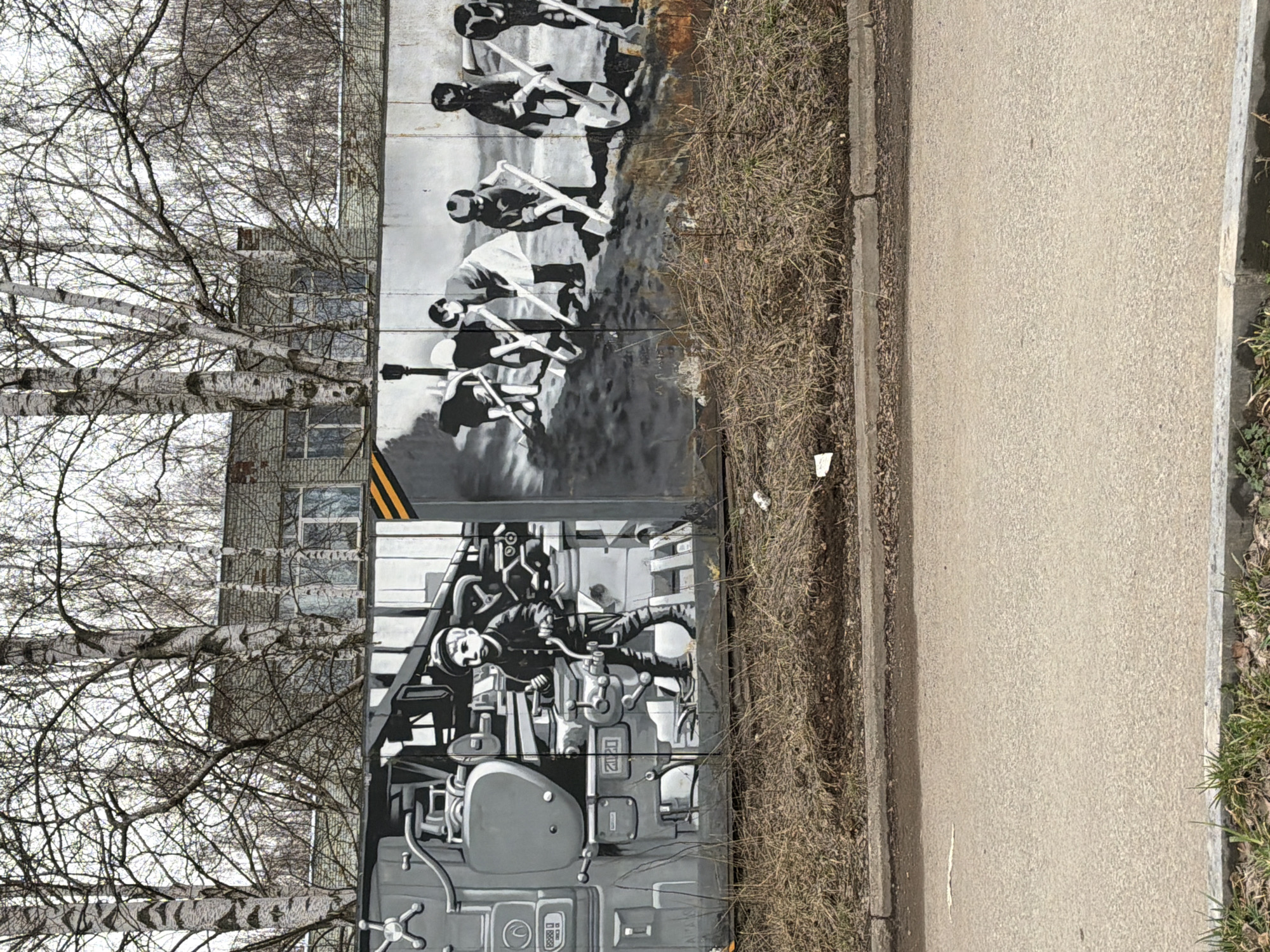 This image, rotated ninety degrees clockwise, captures a scene from Votkinsk, Russia, featuring a long wall adorned with two distinct black-and-white murals. In the foreground, a concrete sidewalk runs parallel to the wall, bordered by dry, brown grass.
The left-hand mural depicts a stylized individual operating complex machinery, possibly a control panel or an industrial engine, characterized by numerous levers, dials, and pipes. The person, rendered with a thoughtful expression, appears deeply engaged in their task.
To the right, a second mural portrays a group of figures, likely a band, playing string instruments resembling guitars or bass guitars. Their poses suggest active performance, with arms raised and instruments held dynamically. The artistic style of both murals is monochromatic, utilizing shades of grey, black, and white to create depth and contrast.
Behind the mural wall, several tall, bare birch trees with characteristic white bark and dark markings stand prominently against a bright, possibly overcast, sky. Through the branches, parts of a multi-story building with visible windows can be seen, its facade appearing textured. The absence of leaves on the trees and the dry grass indicate a cold season, likely late autumn, winter, or early spring. The lighting suggests natural daylight. No legible text is visible within the image.