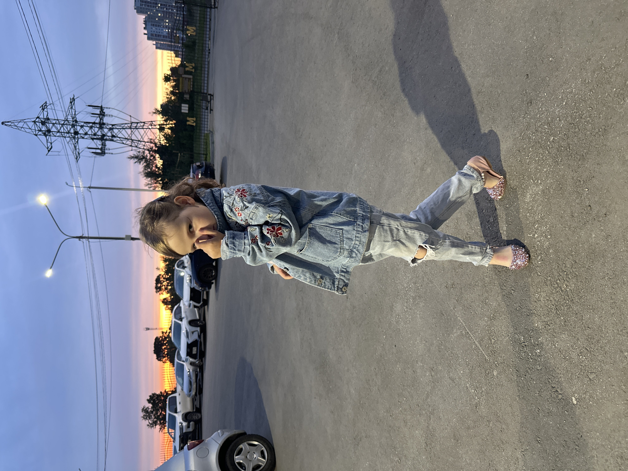 A full-length outdoor photograph captures a young girl standing on a paved surface at sunset in Khabarovsk, Russia. The image is presented vertically, featuring the girl in the foreground with an urban landscape in the background.
The primary subject is a child, appearing to be a pre-teen, with light brown hair tied back. She is dressed in a light blue denim jacket with red floral embroidery, distressed light blue jeans, and sparkly pink ballet flats. She poses with her left hand raised near her chin or mouth, looking towards the camera with a neutral expression.
The setting is an urban or suburban area during dusk. The sky displays a beautiful gradient of deep blue transitioning into purple, pink, and orange near the horizon, indicative of a sunset. A large electricity pylon with power lines dominates the left side of the background. To the right, multi-story apartment buildings are visible.
A row of parked cars, including white, silver, and dark blue vehicles, lines what appears to be a parking lot or street behind the girl. A street light illuminates the scene, and long shadows are cast across the asphalt ground from the low sun. On a fence in the mid-ground, the letter "M" is visibly repeated. The atmosphere is calm, lit by the merging light of the setting sun and artificial lamps.