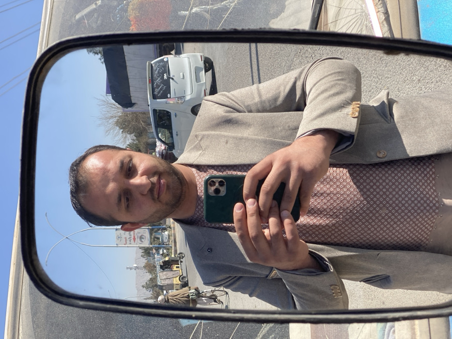 A man in a tan suit and patterned shirt takes a selfie in a side view mirror. The reflection shows a street scene with a white car, other vehicles, and a street sign. In the background are buildings and mountains under a clear blue sky. The mirror's frame is visible, and there are some smudges on the glass. The man holds a smartphone with a multi-camera setup in his hands.