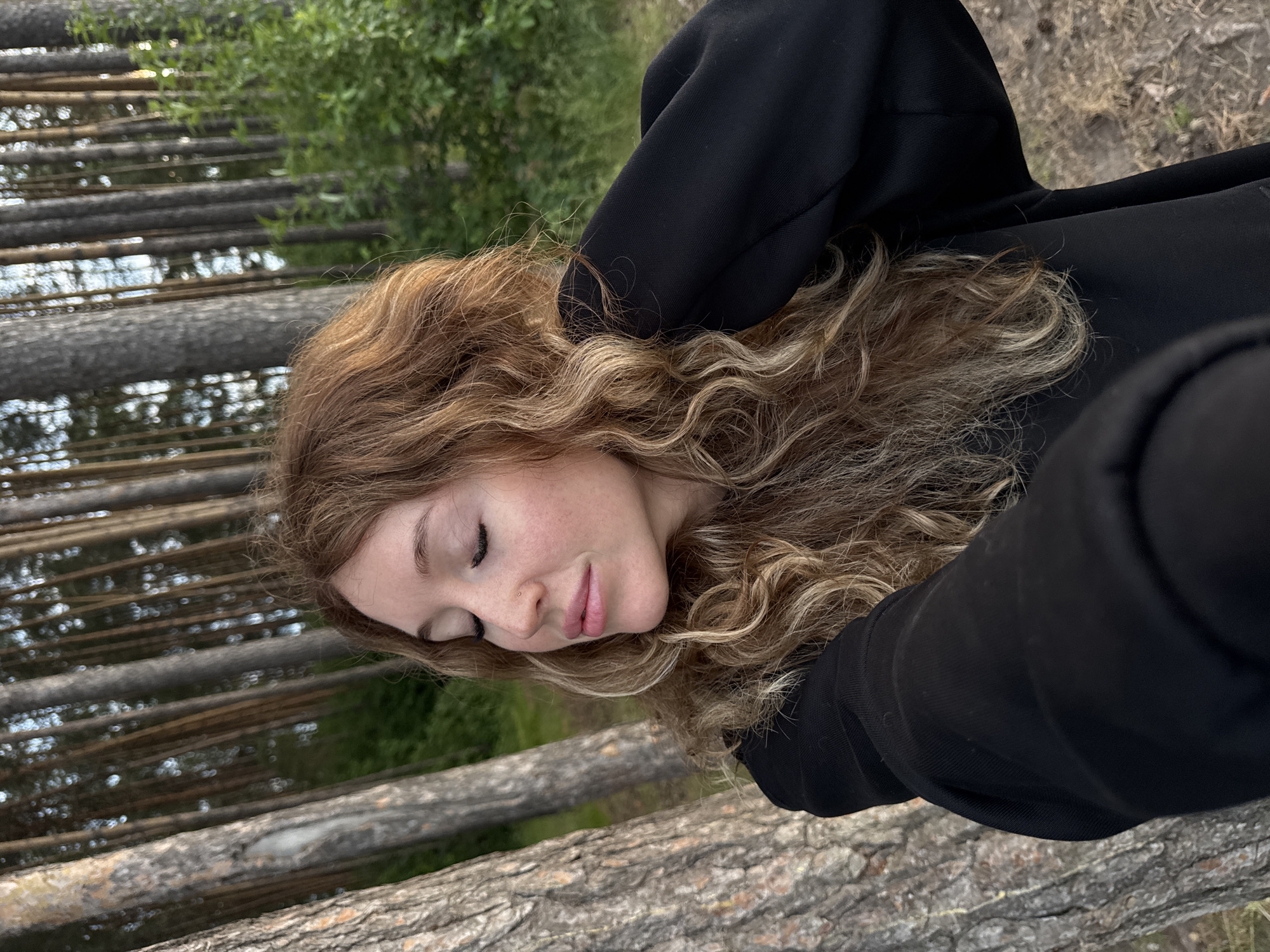 A young woman with long, curly auburn hair is captured in a medium shot, outdoors in a forest setting. She is wearing a black hoodie and has her eyes closed, with her head tilted slightly back and her hand raised to her hair. The background consists of tall, slender trees with rough bark, and dense green foliage. The ground is covered with dry grass and dirt. The lighting suggests it is either early morning or late afternoon.