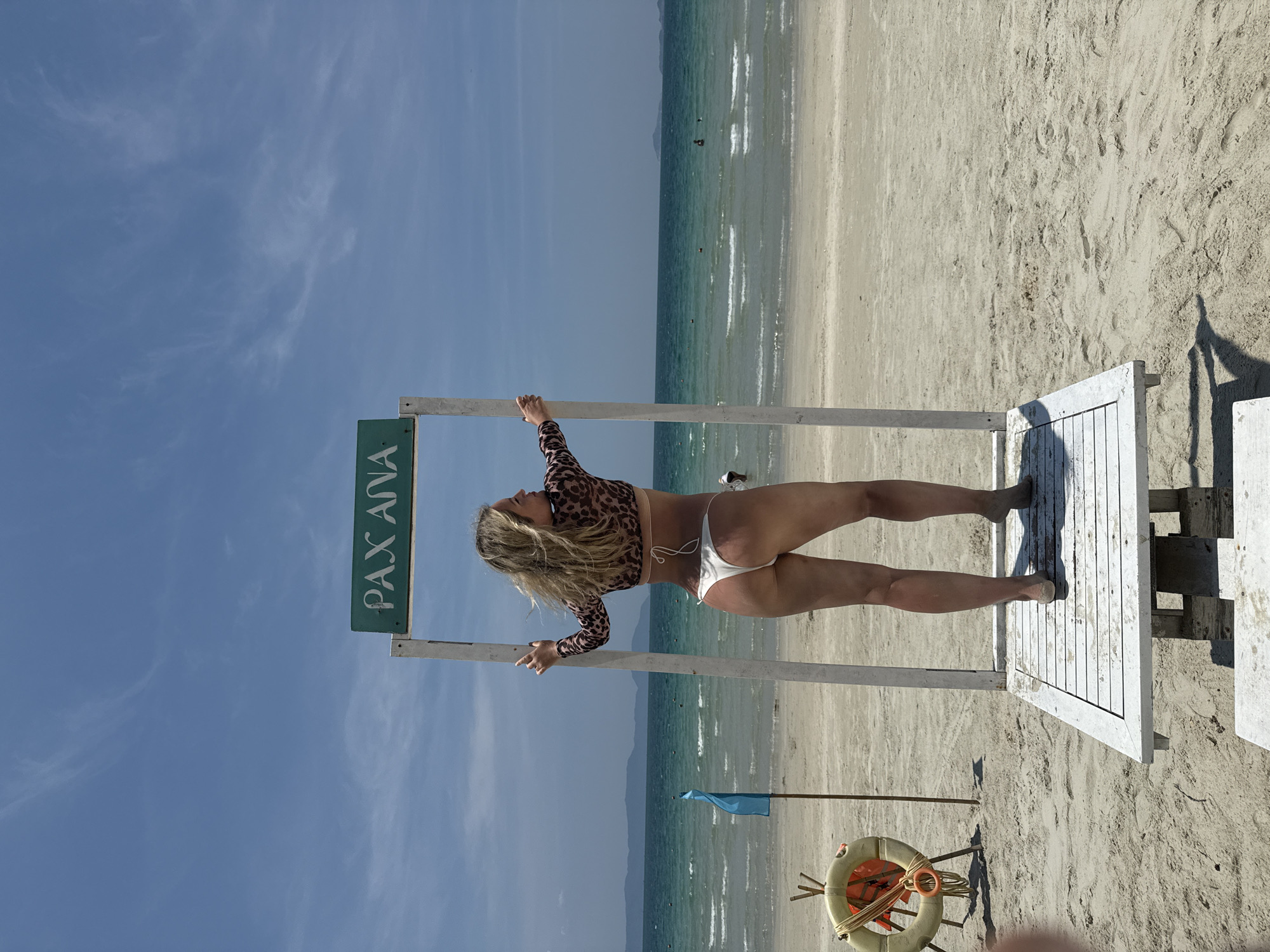 The image depicts a woman in a leopard print top and white bikini bottom, standing on a white wooden platform that is part of a structure with a sign reading "PAX ANA" (oriented vertically). She is holding onto the structure with both hands. The structure is positioned on a sandy beach. In the background is the ocean and a blue sky with scattered clouds. A life preserver and a blue flag are situated near the base of the structure. The location is Dong Ninh Hoa Ward, Vietnam.
