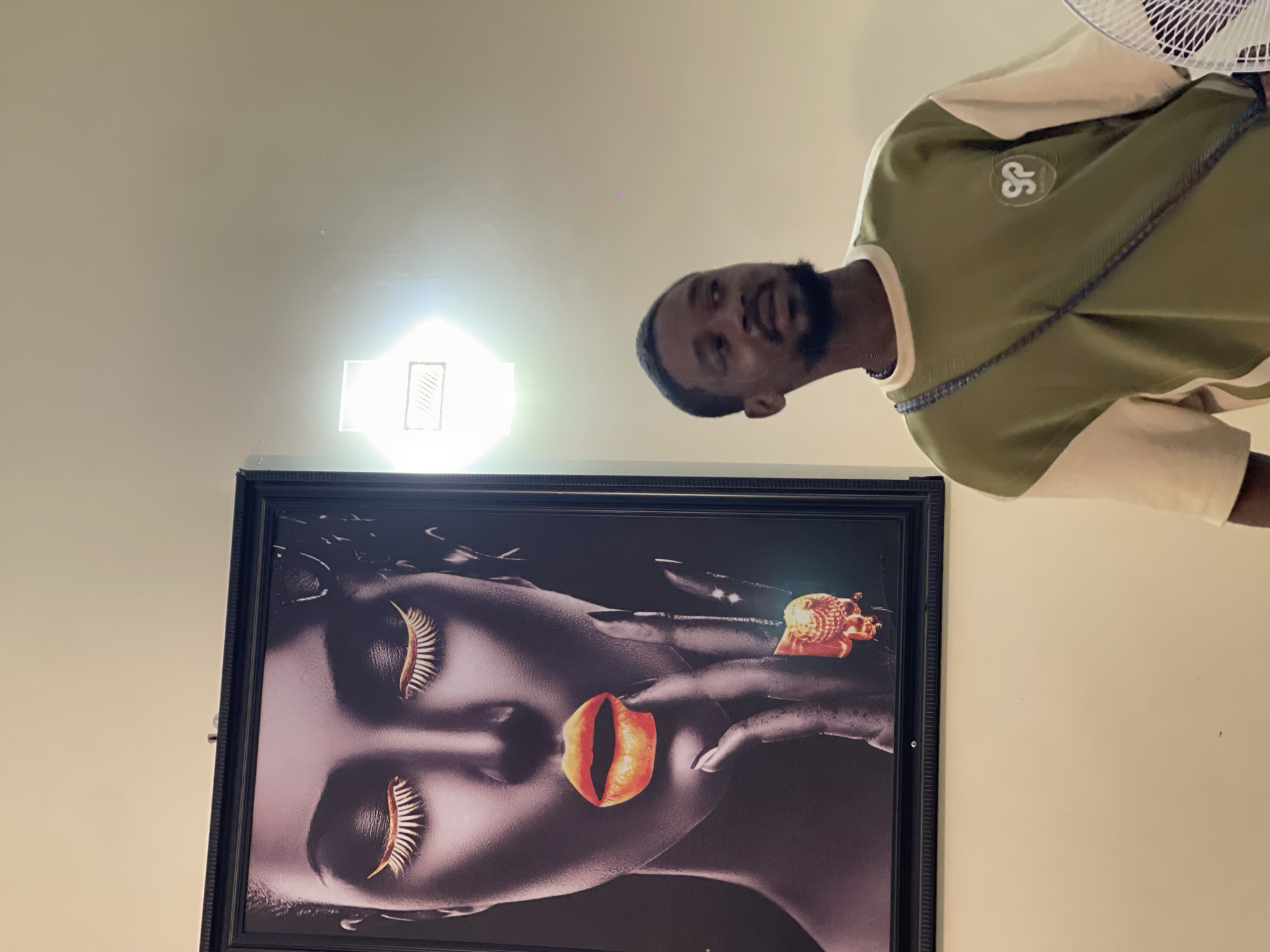 This indoor image, likely taken in Gwarinpa, Nigeria, is oriented vertically and shows a man standing in front of a framed artwork. The man, a young adult with dark skin and a beard, looks directly up at the camera with a neutral to slightly smiling expression. He is wearing an olive green and beige long-sleeved shirt with white stripes on the sleeves and a circular white patch featuring a distinctive "SP" logo on the chest. A dark necklace is visible around his neck.
Behind and to the left of the man, a large, black-framed picture is mounted on a light-colored wall. The artwork depicts a stylized profile of a woman's face, rendered in dark, grayscale tones. She has closed eyes with striking, golden, feather-like eyelashes. Her lips are a vibrant, prominent red-orange, appearing slightly open and possibly resembling a flower petal or a unique fruit. A hand with dark, manicured nails is positioned near her face, holding a intricate, golden, snail-like object near her mouth.
The background consists of plain, light-colored walls and a ceiling. A bright, rectangular ceiling light fixture is prominently visible, casting a strong glare in the upper left quadrant of the image. Part of a white fan, likely a ceiling fan, is visible in the top right corner of the original image, partially cropped out in some hints. The overall lighting is bright and artificial, suggesting an indoor setting without a clear indication of the time of day.
