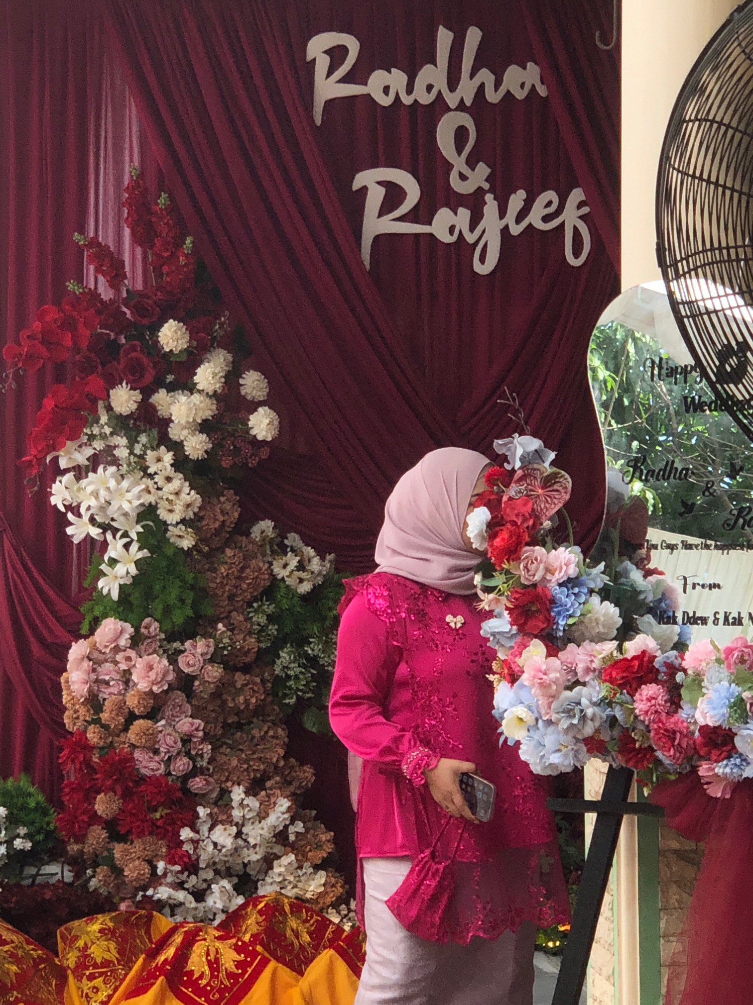The image depicts a wedding celebration with floral decorations and a backdrop of maroon drapery. A person wearing a pink hijab and a sequined magenta top stands beside a large floral arrangement. In the background, white cursive lettering on the drapery reads "Radha & Rajcef". A circular mirror with text written on it is also visible. The scene is set outdoors, with greenery and sunlight suggesting a daytime event, possibly in Lambadalhok, Indonesia, as indicated.