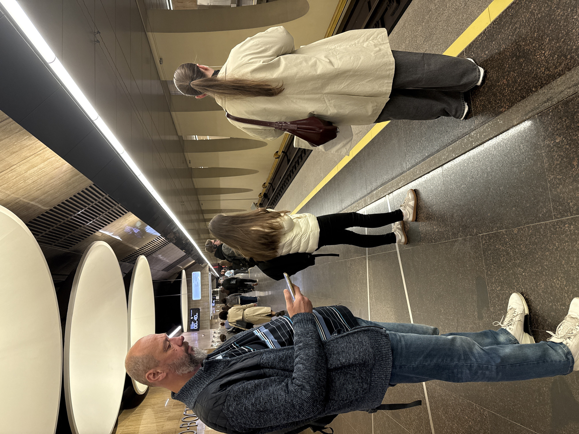 The image captures a scene in a subway station in Moscow, Russia. The station features a modern design with large, illuminated circular panels lining the platform wall. Several people are waiting for a train. One man is looking at his phone. Other people are standing further away from the camera, closer to the platform edge. A person wearing a tan jacket with a brown purse and another person wearing black leggings are standing close to the edge of the platform. The platform is marked with a yellow line.