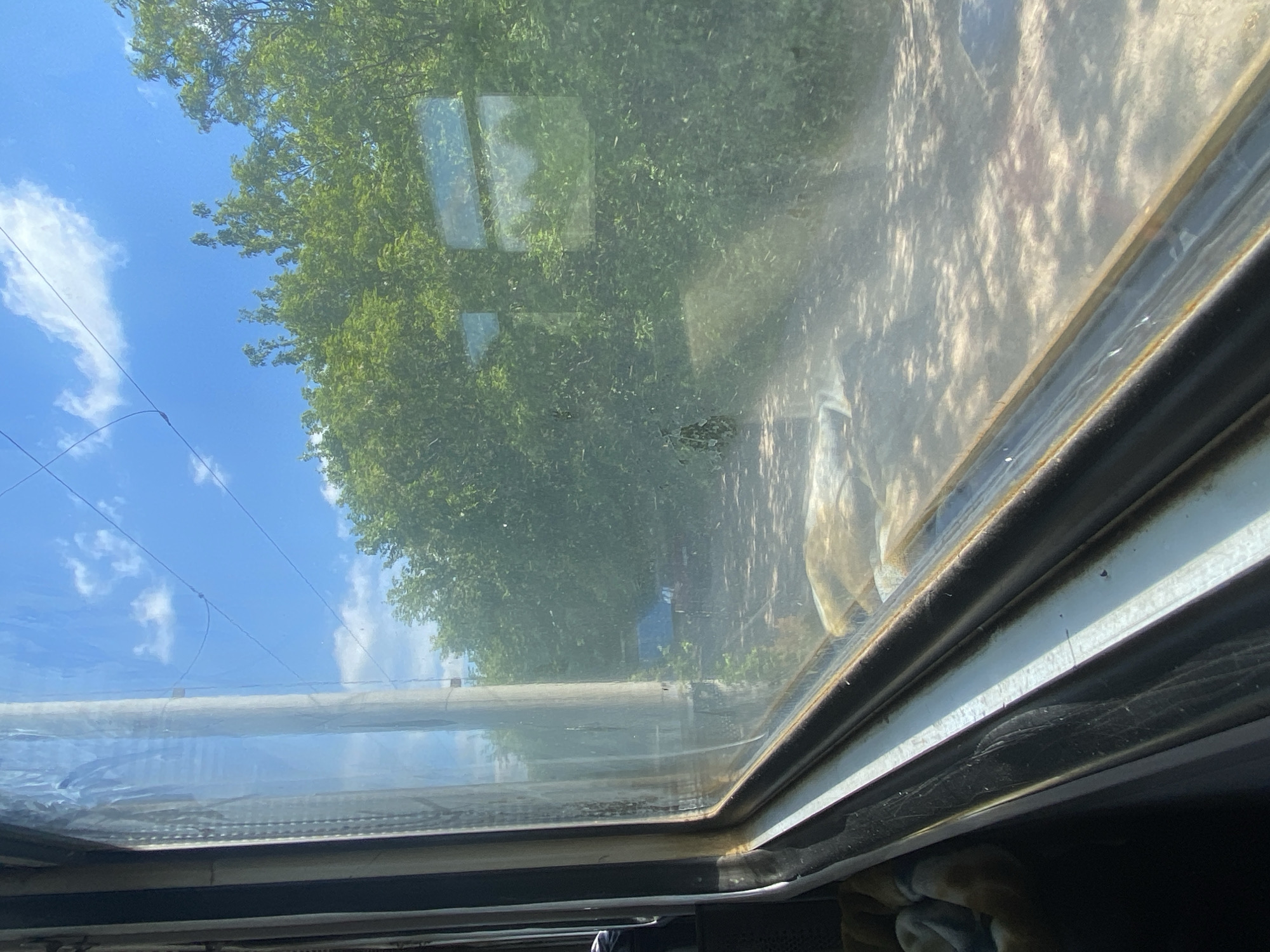 The image shows a view through a dirty window. Outside, there are trees and a blue sky with some clouds. A street is visible in the reflection. The location is Matiashivka, Ukraine, although there are no identifying landmarks to confirm the location.