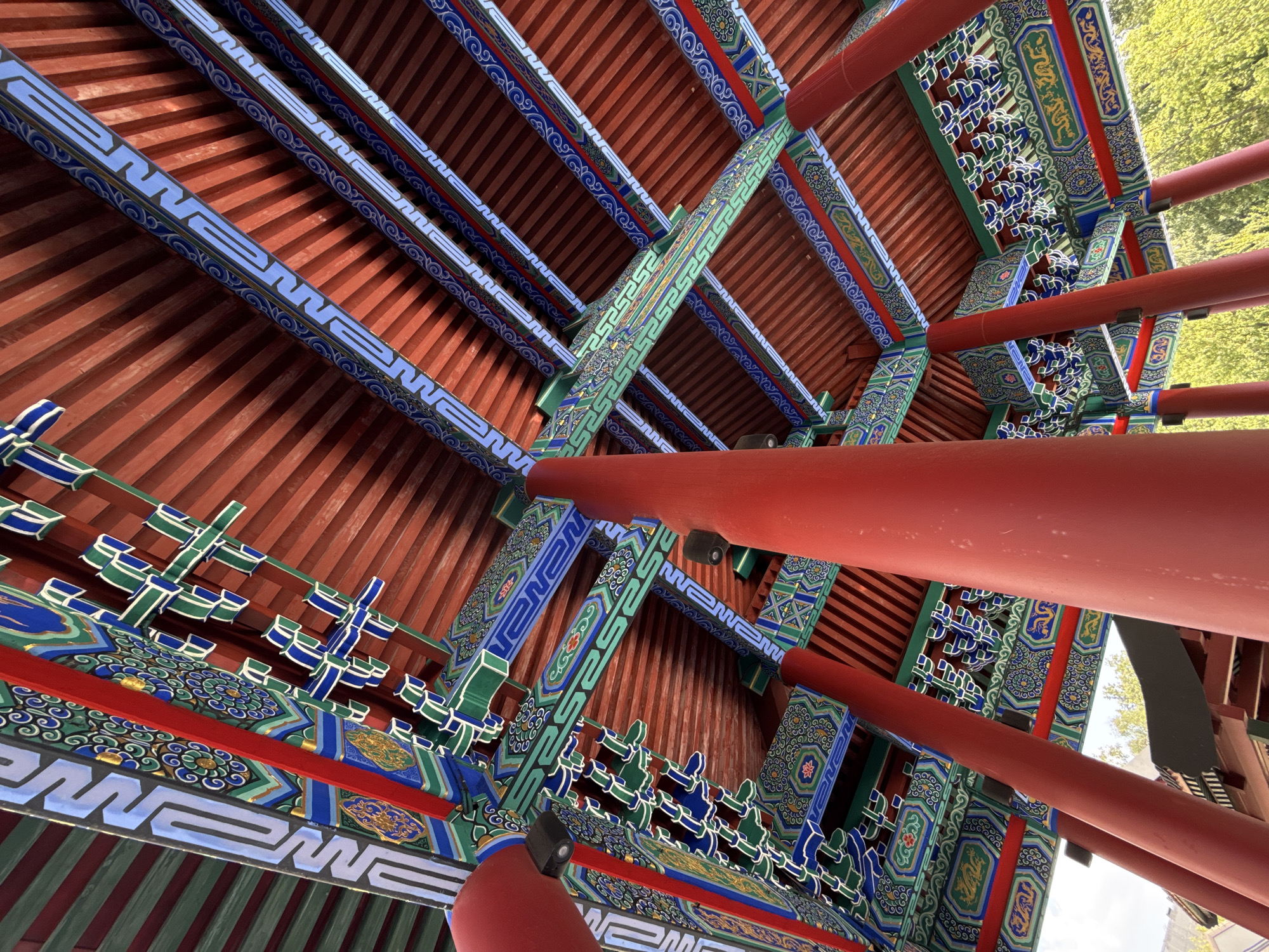 This image shows an upward view of a brightly colored, ornate roof structure of a Chinese-style pavilion. The roof is supported by several thick, red cylindrical pillars. The ceiling is constructed of parallel wooden planks painted red, and intricate decorative beams painted in red, green, blue, and gold adorn the underside of the roof. The beams feature detailed geometric patterns and motifs, including stylized clouds, swirling patterns, and what appear to be dragons. The overall impression is one of traditional East Asian architectural style.
This is the roof of the Chinese Pavilion in the Moscow State University Botanical Garden, also known as "Aptekarsky Ogorod".