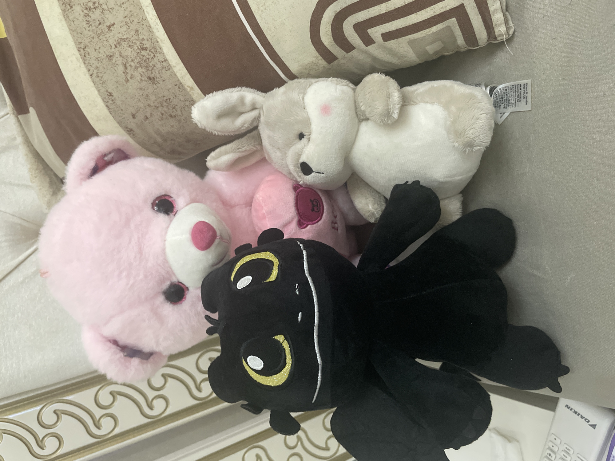 The image displays three plush stuffed animals positioned on a light grey upholstered surface. In the foreground, on the left, is a black dragon plush, identifiable as Toothless from *How to Train Your Dragon*, featuring large yellow eyes with white highlights and a stitched white smile. Behind it, in the center, is a pink bear plush with dark, glittery eyes, a pink nose, and a small dark pink heart-shaped patch on its chest. To the right of the black dragon, and slightly in front of the pink bear, is a light grey and white rabbit plush with small black eyes, a stitched black nose, and pink blush marks on its cheeks. A white fabric tag is visible extending from the rabbit's underside, with partially discernible text including "Oxford UK London".
On the far lower-left of the frame, a white Daikin branded remote control rests on the grey surface. The background on the left features a white panel adorned with gold-colored scrollwork. The right background consists of two cushions: one with alternating brown and cream/white stripes, and another below it exhibiting a brown geometric pattern.
The image contains no visual information to determine or confirm its location in the city of Ihtiman, Bulgaria.