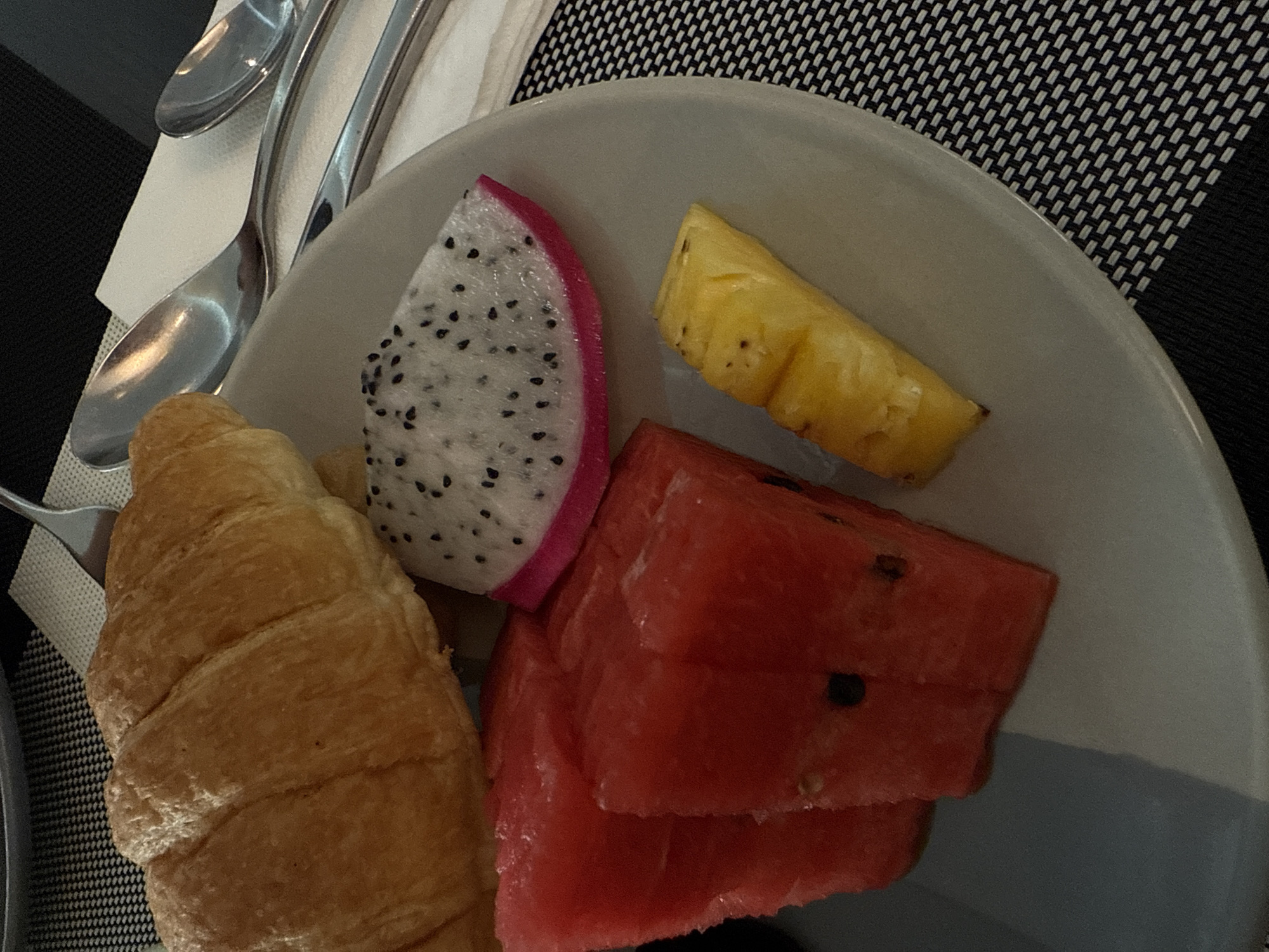 The image presents a top-down view of a breakfast plate. The plate, a muted gray color, contains a croissant, a slice of dragon fruit with a pink rind and black seeds, a slice of pineapple, and several pieces of watermelon. Utensils, including spoons, are visible to the side of the plate. The table is covered with a black and white woven placemat. While it is impossible to determine the location as Rawai, Thailand from the image alone, the food items could be consistent with the cuisine found there. There is no information about any people.