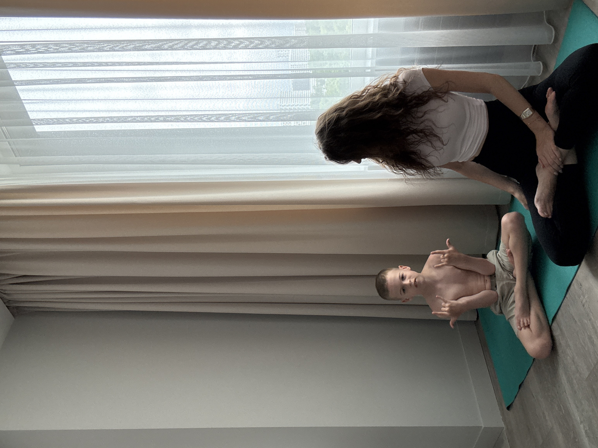 The image shows a woman and a young boy on yoga mats in a room with large windows covered by sheer curtains. The woman, with long, wavy brown hair, is wearing a white top and black leggings. She is in a yoga pose, with one leg bent and crossed over the other, and her hands are holding her foot. She is looking down towards the boy. The boy is shirtless and wearing light-colored shorts. He is also in a yoga pose, with his legs crossed, and he is making a "rock on" hand gesture with both hands. He is looking directly at the camera with a playful expression. The room appears to be well-lit, likely due to the large windows, suggesting it is daytime. The overall mood of the image is relaxed and playful, with the woman engaged in a yoga practice and the boy participating with a sense of fun.