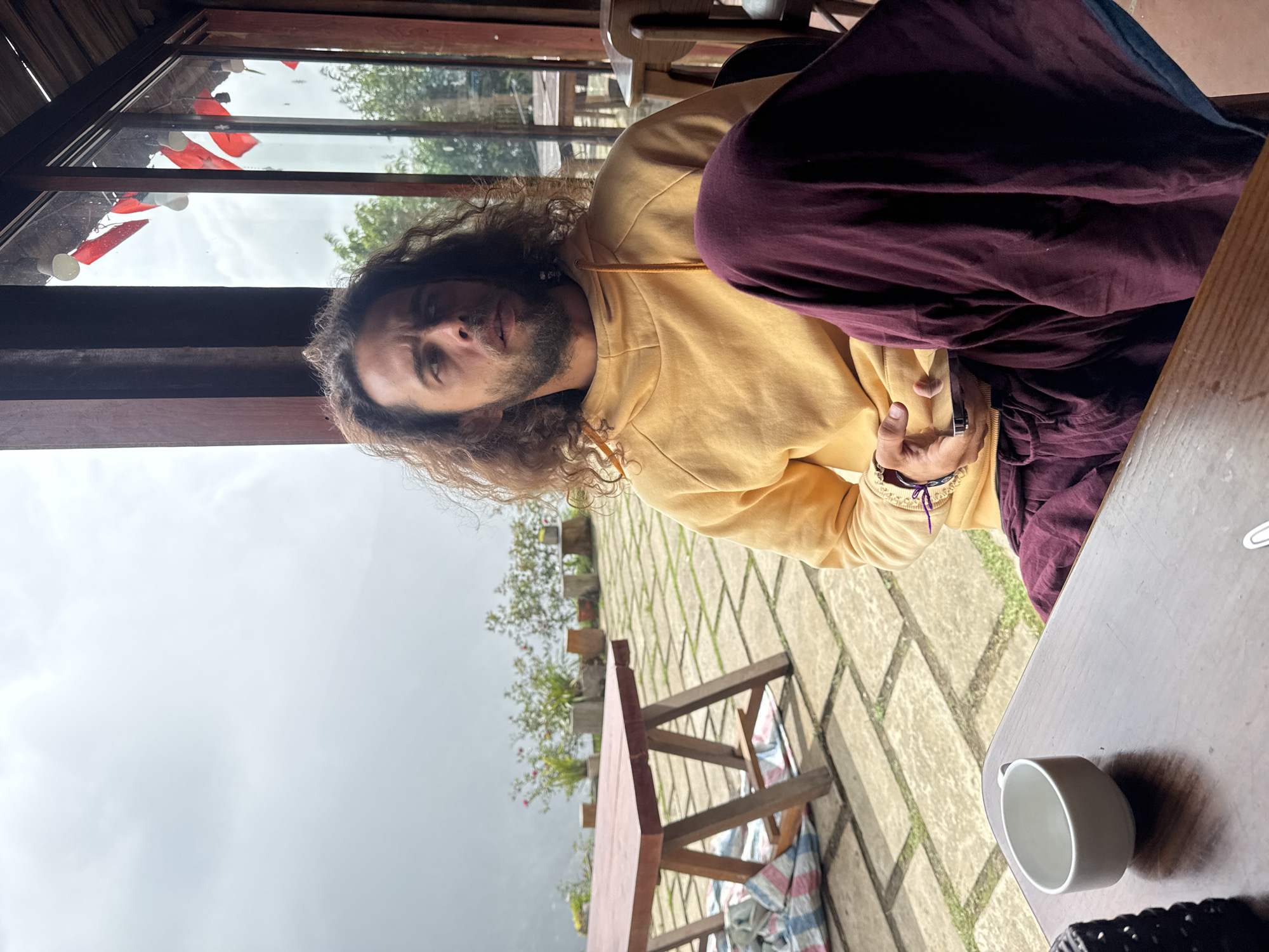 A man with long, curly brown hair and a beard sits at a wooden table inside a cafe with large windows. He is wearing a yellow hooded sweatshirt and dark purple pants, with a piece of dark fabric draped over his lap. In his right hand, he holds a smartphone. To his left, on the table, is a white teacup. Outside the window, a patio with flagstone paving is visible, with potted plants and a wooden picnic table. Beyond the patio, a misty landscape with trees and hills can be seen. The cafe appears to be located in Ta Xua, Vietnam, given the distinctive architecture and natural setting often found in the region, known for its cloud seas.